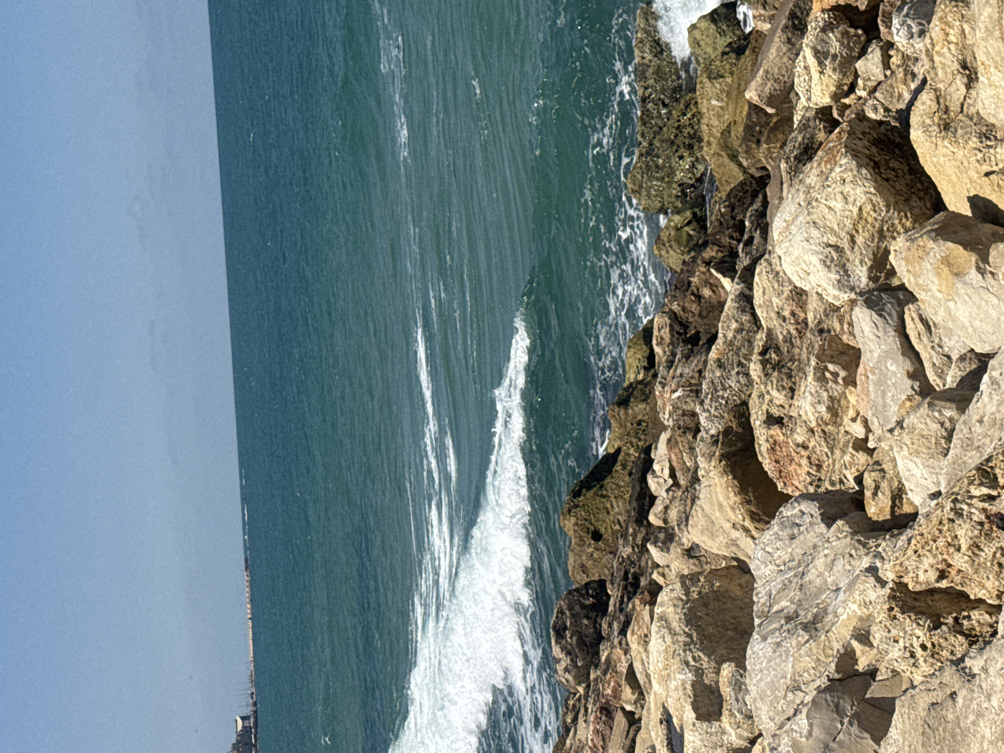The image shows a coastal scene in Tel Aviv, Israel. The sea, with waves crashing, occupies the left portion of the frame. On the right, a rocky embankment forms the edge between the sea and land. In the distance, along the horizon, there is a faint suggestion of buildings, possibly part of the Tel Aviv skyline. The sky is a clear, light blue. No people or specific objects are clearly visible beyond the rocks and sea.