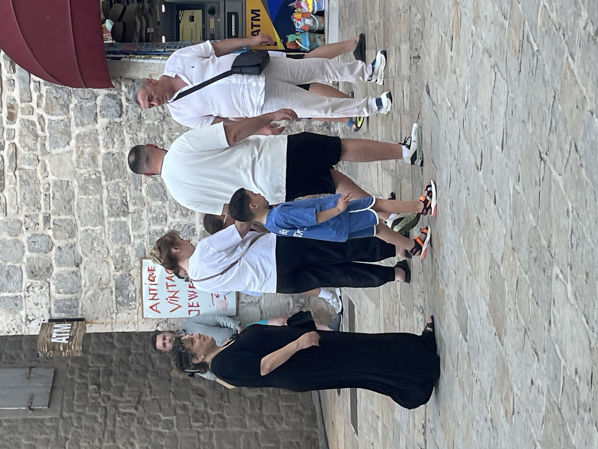 In Budva, Montenegro, on a stone-paved street, a group of people are gathered. A woman in a long black dress walks from left to right. To her right, a man in a light blue shirt stands behind a sign that reads "ATM". Further right, a woman in a white t-shirt and black pants looks down, and next to her, a man in a white t-shirt and black shorts is partially visible. A young boy in a blue t-shirt and shorts walks in front of the man in white. Another man in a white linen shirt and light-colored pants stands to the right, facing the man in the white t-shirt. Behind him, an ATM machine is visible. In the background, a stone wall and a red awning are present.