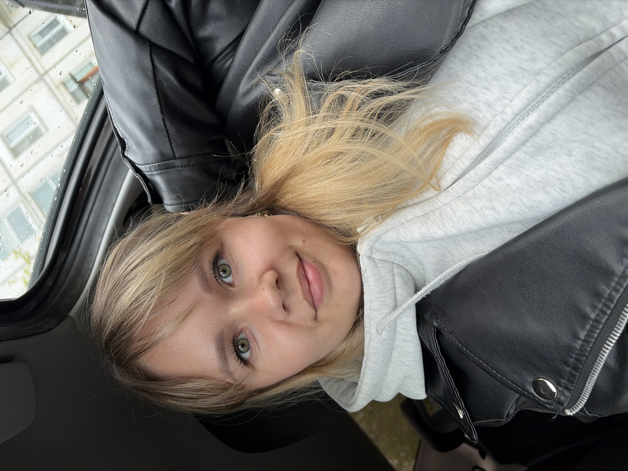 A young woman with blonde hair and green eyes is sitting in a car. She is wearing a light gray hoodie and a black leather jacket. It appears to be a rainy day, as there are water droplets on the car window, behind which, a building with white windows is visible. No other information about the location in Chaikovsky, Russia is available from the image.