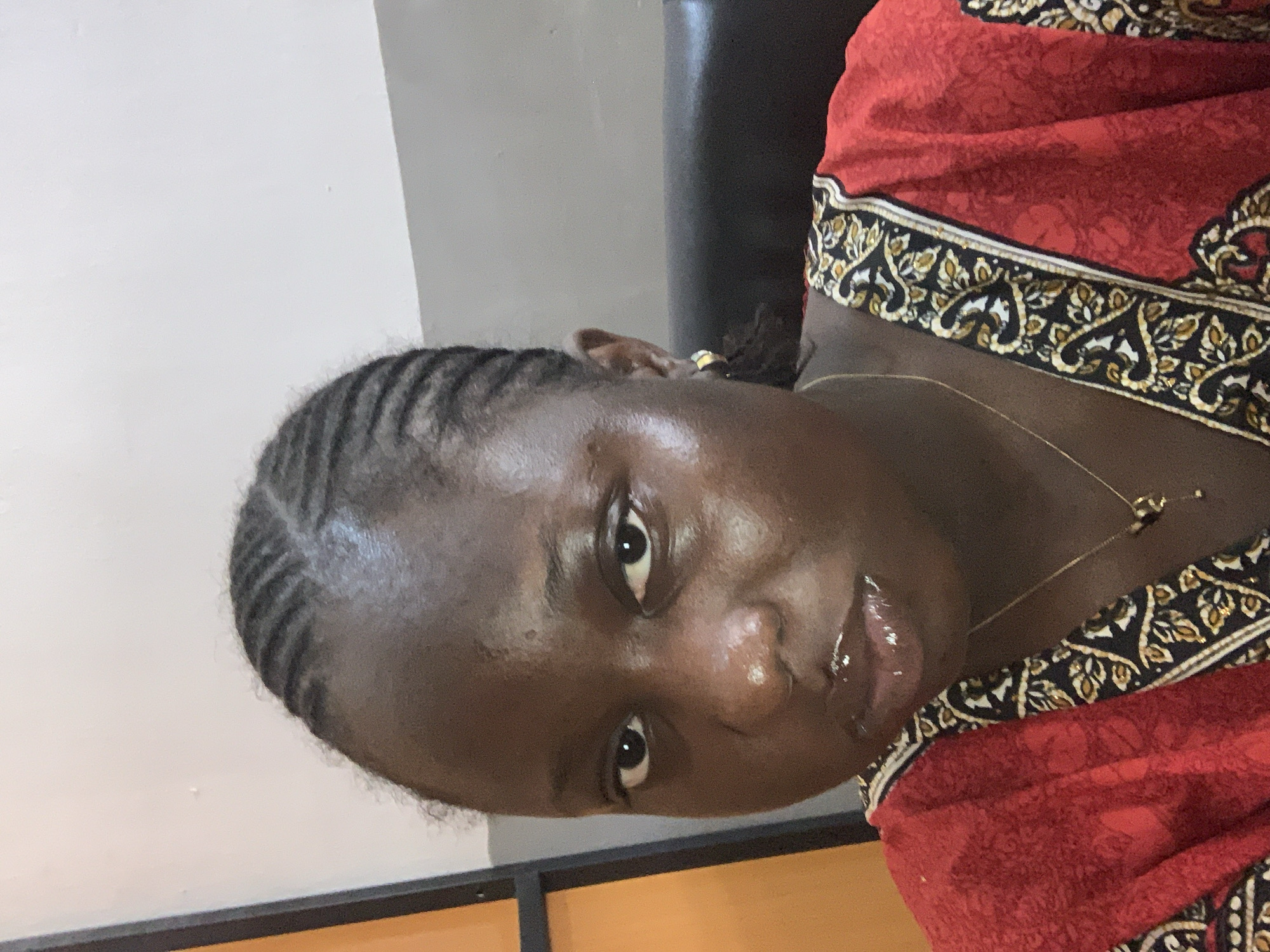 A medium shot portrait shows a woman with dark skin and braided hair. She is wearing a red top with a black and gold patterned trim. Her eyes are open and looking forward, and her lips are glossy. A gold necklace with a small pendant rests on her chest. The background is out of focus and appears to be an indoor setting with a white wall and a dark surface behind her. The lighting is bright, highlighting the shine on her face.