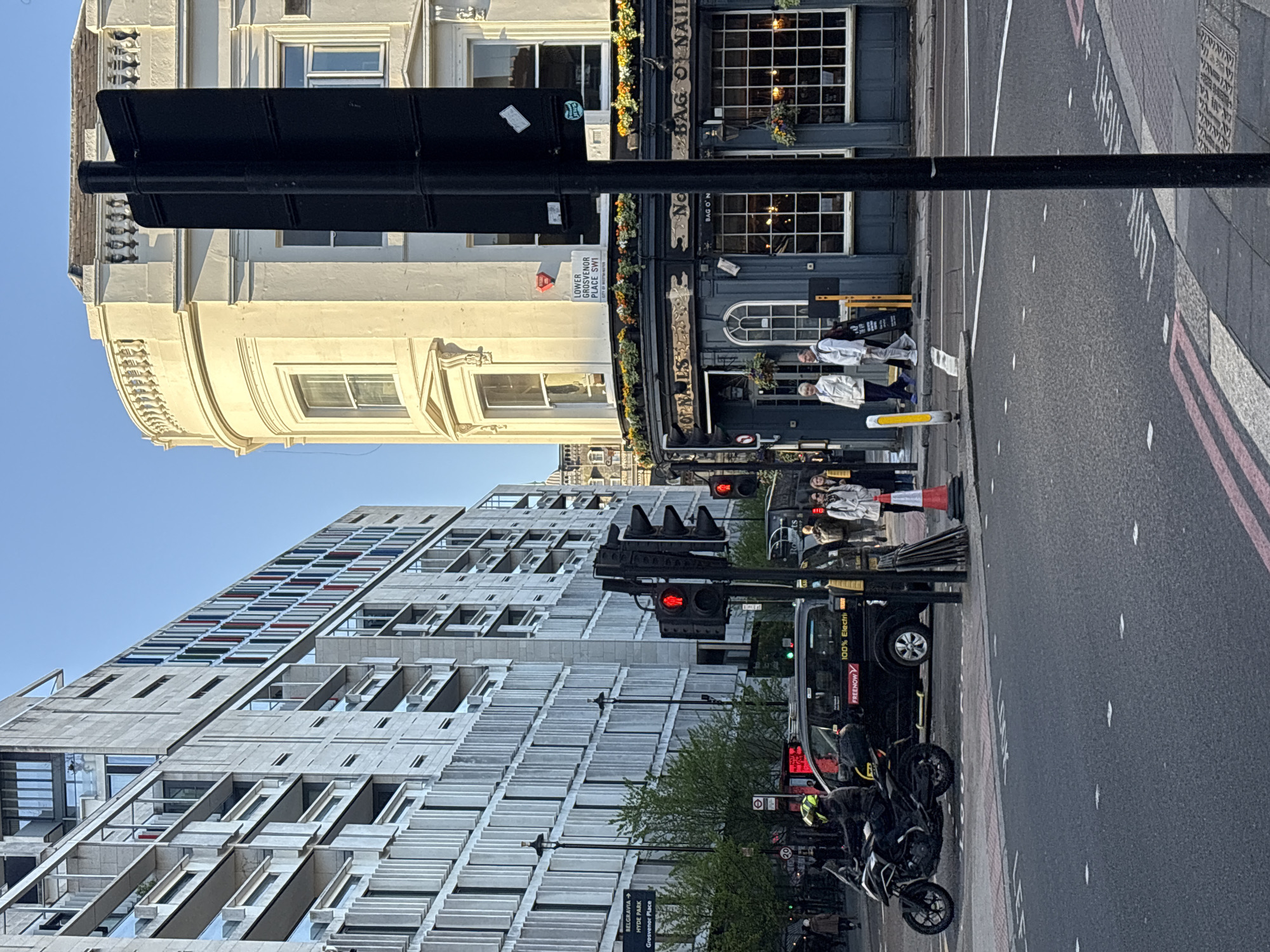 The image captures a street scene in London, United Kingdom. A multi-story building with distinctive architectural features stands on one side of the street, while a traditional pub-style building, "Bag o' Nails," occupies the opposite side. Street signage indicates the location as Lower Grosvenor Place, SW1, near Belgravia, Hyde Park, and Grosvenor Place. Traffic lights are visible, and a black electric taxi with the "FREENOW" logo is parked on the road. Pedestrians are present, and motorcycles are parked nearby. The overall environment suggests an urban setting in London.
