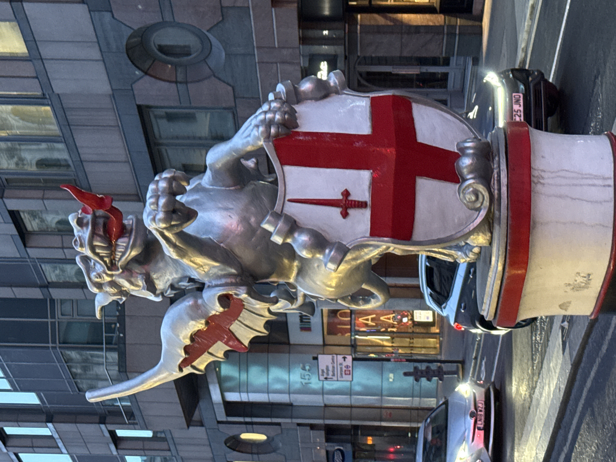 The image shows a griffin statue holding a shield, likely located in the City of London, United Kingdom. The griffin is silver, with red accents on its wings and the flames coming from its head. The shield it holds is white with a red cross and a smaller red sword in the lower center. The statue sits atop a white column with a red band around the top. In the background, there is a modern building with reflective windows. Street signs indicate the direction of the Barbican Centre and Liverpool Street. Cars are visible on the street, with license plates LN18 WZJ and C25 JNO.