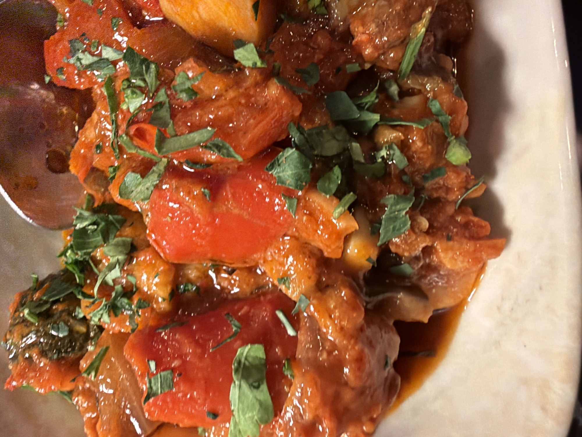 A close-up shot presents a plate of what appears to be stew or a similar cooked dish, possibly of a Mediterranean or Middle Eastern style. The main components include chunks of tomato, possibly other vegetables, and what could be meat or a meat substitute, all covered in a rich, glossy sauce. Fresh green herbs, perhaps parsley or cilantro, are scattered across the top as garnish. A spoon is partially visible on the left. Given the image context, the location of this image is unlikely to be determined.