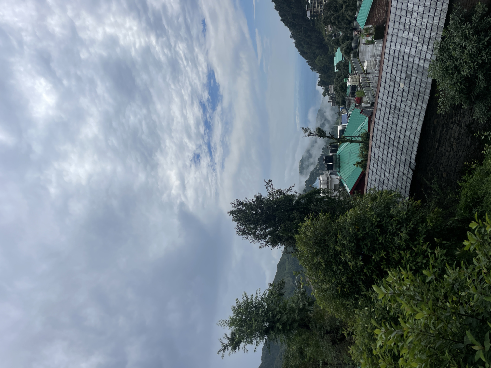 The image depicts a view of a hillside town with a cloudy sky overhead. In the foreground, there is dense green foliage. Behind the foliage, a building with a gray, slate-tiled roof and stone walls is visible. Further into the scene, the town extends down the hillside, characterized by numerous buildings with vibrant green roofs. Some of these buildings have water tanks on their flat rooftops. The distant landscape is characterized by rolling hills covered in trees, with a hazy atmosphere obscuring the furthest views. The sky is filled with layered clouds, with patches of blue sky visible in the upper right portion of the image. The scene suggests a mountainous or hilly region in Bhagsu, India, likely taken from an elevated viewpoint looking down into the town and towards the surrounding natural landscape. There are no people or animals visible in the image.