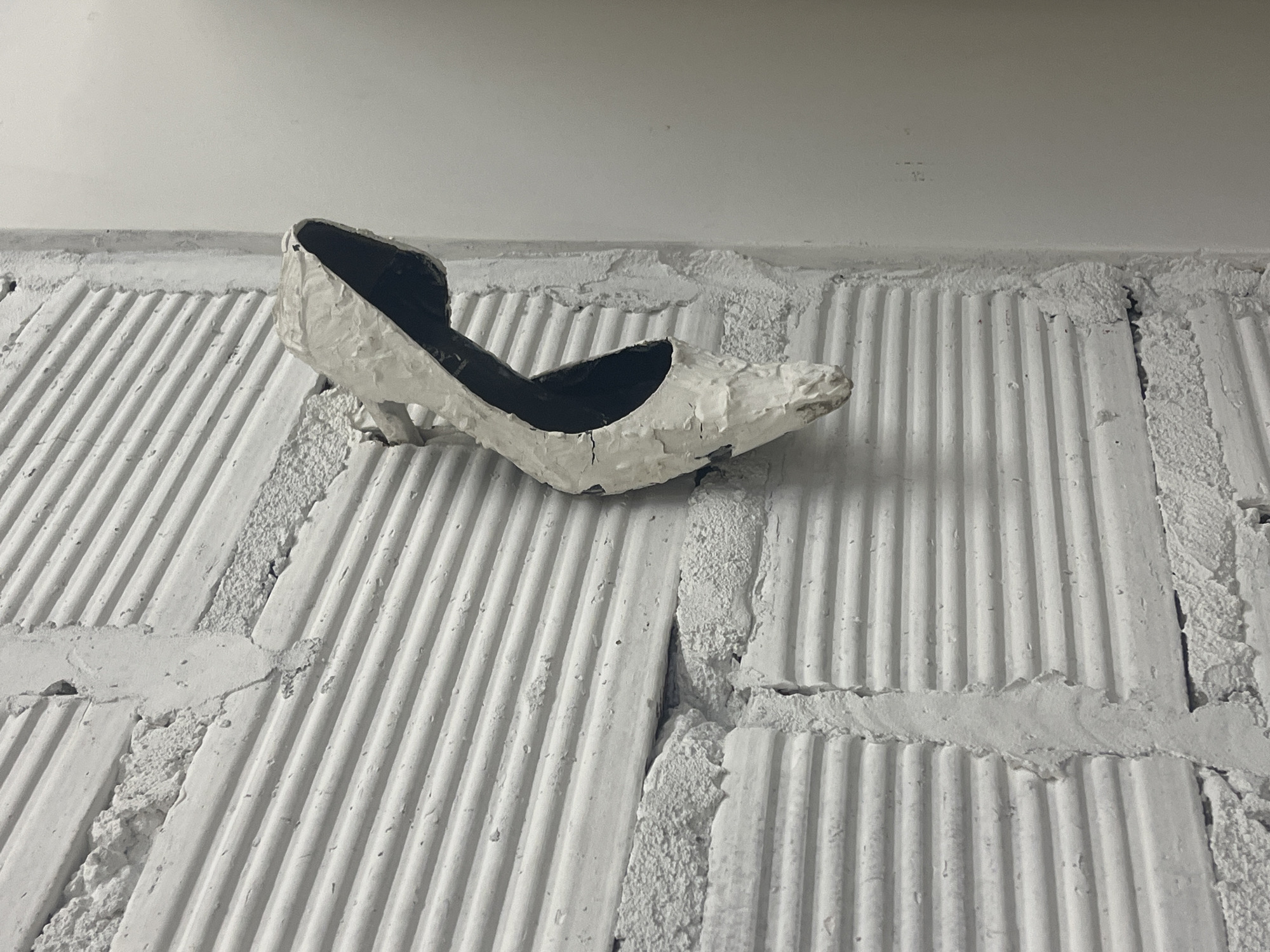 A white, damaged high-heeled shoe sits atop a section of painted white brickwork. The shoe has a dark interior. The brickwork has a ridged texture and is laid in a block pattern with rough, irregular edges between the blocks. The scene appears to be indoors, near a ceiling. The specific location in Buenos Aires, Argentina, cannot be determined from the visual information provided.