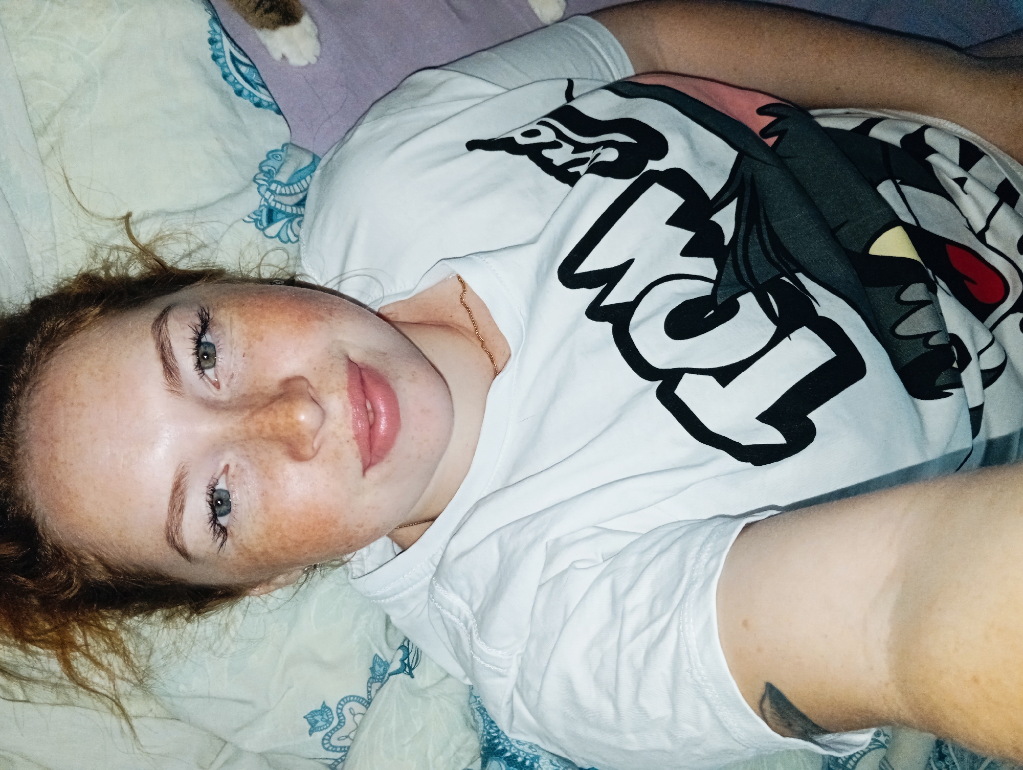 A young woman with red hair and freckles is lying on a bed. She is wearing a white t-shirt with a cartoon graphic on it. Part of a cat's paw is visible in the upper left corner. The bed has a light blue and white patterned comforter. The location is in Yurgamysh, Russia.