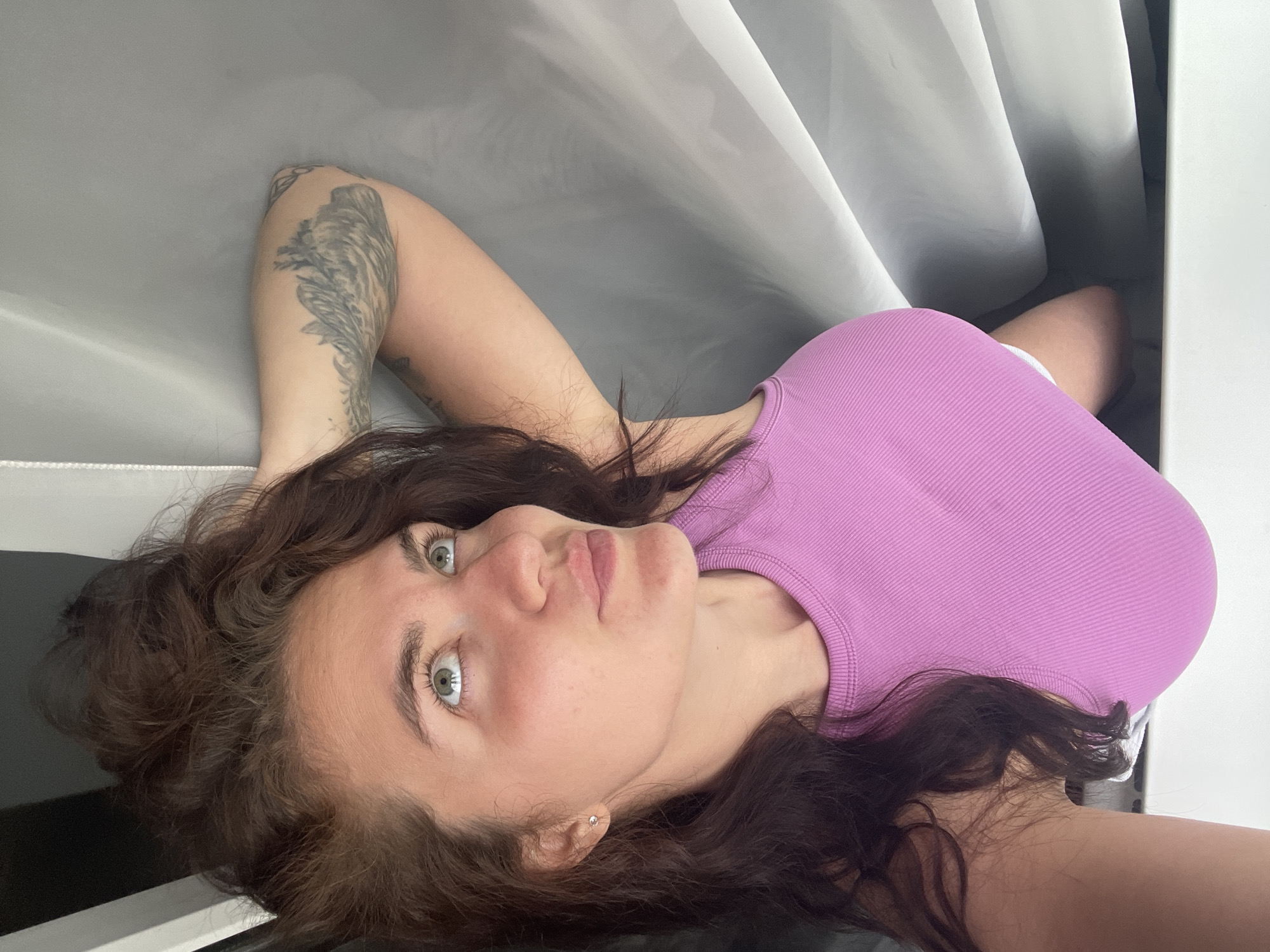 A woman with brown hair and green eyes lies down in a confined space. She wears a light purple ribbed top. Her left arm is raised, revealing a tattoo. She is positioned near what appears to be a curtain or sheet. The location information is not present in the image.