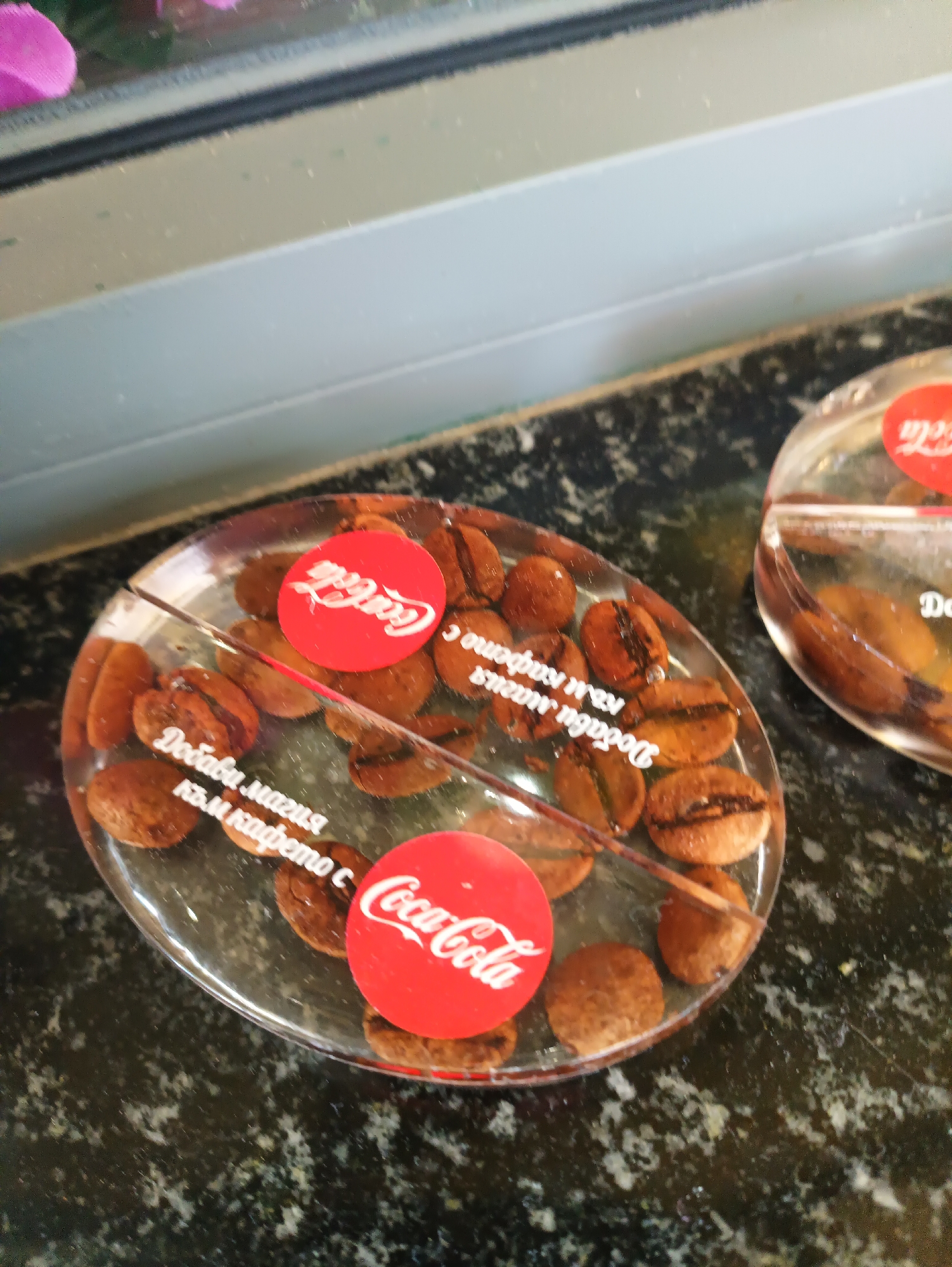 The image shows two clear plastic containers, each divided into two sections. Inside each section are coffee beans. Each container has a Coca-Cola logo sticker on each of the two sections. The text "Добави магия към кафето с Coca-Cola" (which translates to "Add magic to coffee with Coca-Cola") is printed around the edge of the sections, partially obscured by the coffee beans. The containers sit on a dark speckled surface. The image suggests a promotional item or display related to Coca-Cola and coffee. It may be located in Karnobat, Bulgaria.