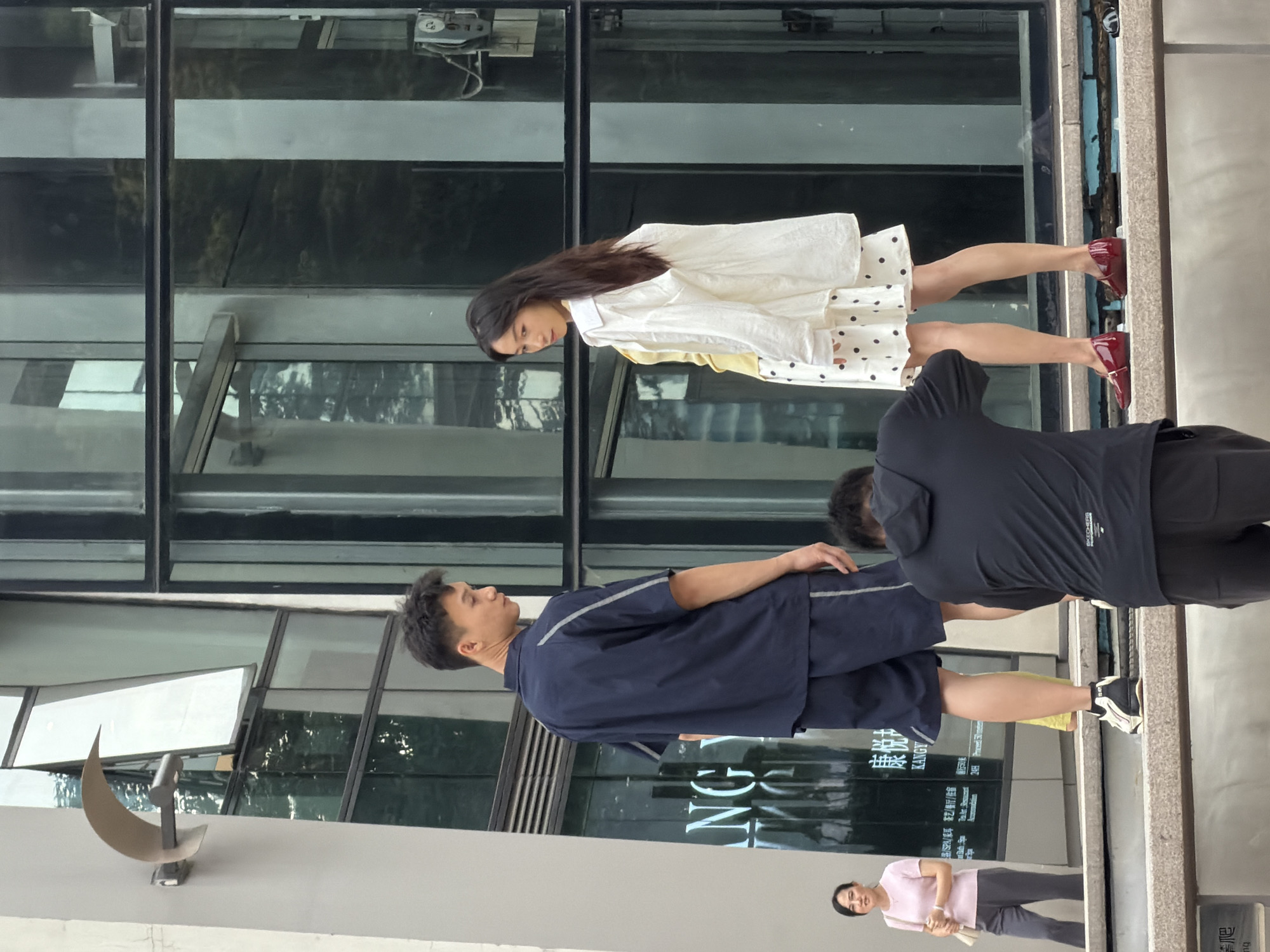 In Yuzhong District, China, a group of individuals are captured in an outdoor urban setting with a modern glass-fronted building in the background. The scene appears to be a street or a public plaza, possibly part of a commercial or shopping area. In the mid-ground, a man and a woman are standing, positioned on a raised platform or steps. The man is wearing a dark blue short-sleeved shirt and matching shorts. The woman is dressed in a white shirt worn open over a yellow top, and a white skirt with black polka dots. She is also wearing dark red flat shoes. Another individual, seen from the back and wearing a black hoodie and dark pants, is crouching in the foreground, seemingly taking a photograph or video of the man and woman with a camera. To the left of the frame, further in the background and on a lower level, a woman in a pink t-shirt and grey pants is walking away from the camera, carrying a light-colored bag. The building behind the subjects features large glass panels, reflecting the surrounding environment and sky. A metallic sculpture, curved and abstract, is mounted on the building's facade to the left. The overall atmosphere suggests a casual daytime activity, possibly a fashion shoot or a moment of street photography.