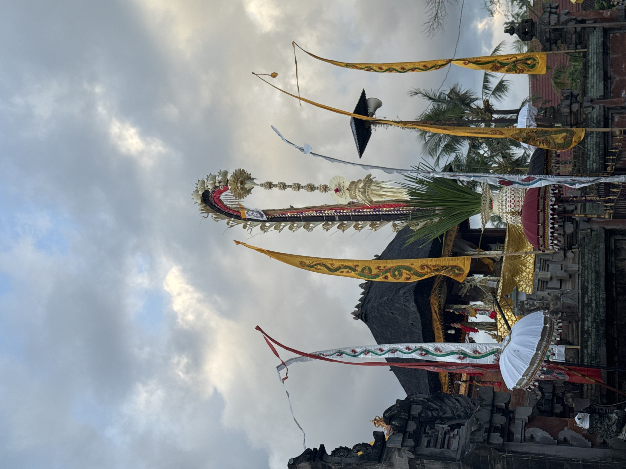 The image depicts a view of a temple complex in Blahbatuh, Indonesia, under a cloudy sky. In the foreground, several traditional Balinese flags are visible, including tall, yellow flags with green serpent-like designs, and a white flag adorned with green and red floral patterns. A white ceremonial umbrella with gold trim is positioned to the left, and a red umbrella is partially visible on the right. Behind these, a multi-tiered structure with a dark thatched roof, adorned with gold accents and intricate carvings, forms the main temple building. Religious effigies are visible within the open-air sanctuary. Stone sculptures and decorative fencing are also present. Palm trees can be seen in the background, suggesting a tropical environment. The overall scene suggests a cultural or religious ceremony is underway or has recently concluded.