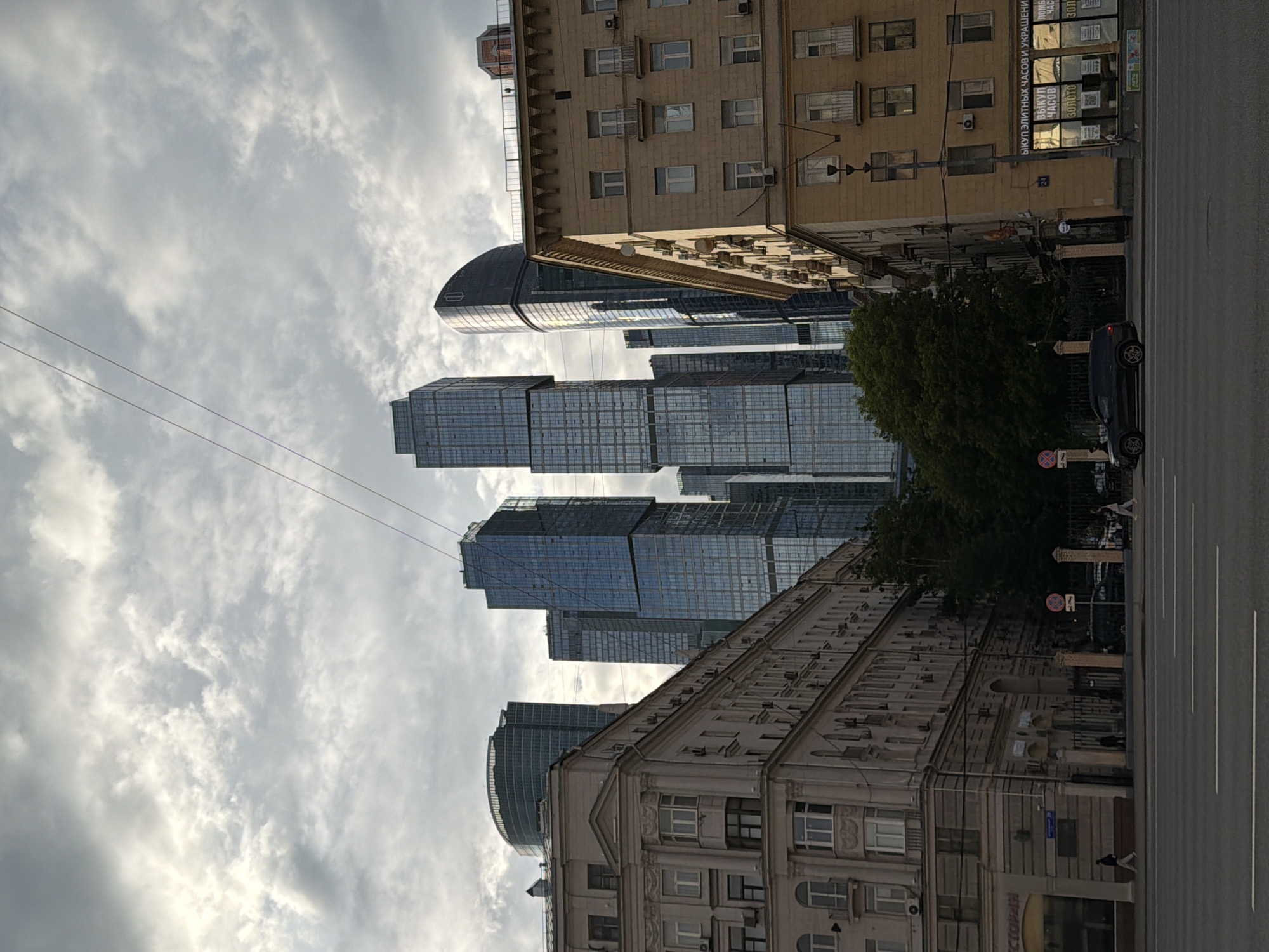 This is a ground-level, medium shot photograph taken on a cloudy day, looking up at a mix of old and new architecture in Moscow, Russia. The foreground features a wide street with visible lane markings and a few pedestrians walking on the sidewalk. On the right side of the street, a light brown, older apartment building with multiple windows and balconies dominates the frame. Some of the windows have air conditioning units installed. The building's facade shows signs of age and wear. On the left, a similar older building, painted a lighter beige, is visible with more detailed architectural features, including arched windows and decorative elements.
Behind these older buildings, a cluster of modern skyscrapers pierces the sky. The most prominent are two tall, glass-facade skyscrapers with a distinctive stacked design, reflecting the cloudy sky. To their left is a rounded, dark-colored skyscraper, and other smaller, contemporary office buildings are also visible. A dense green tree is nestled between the older buildings and the modern skyscrapers, providing a stark contrast in textures and colors. Two thin wires, likely power lines or communication cables, stretch diagonally across the sky. A dark-colored SUV is parked on the street in front of the older buildings on the right. Signs are visible on the storefronts of the older buildings, with some text in Cyrillic, indicating commercial establishments. The overall impression is a view of the Moscow skyline where historical urban fabric meets modern development.
