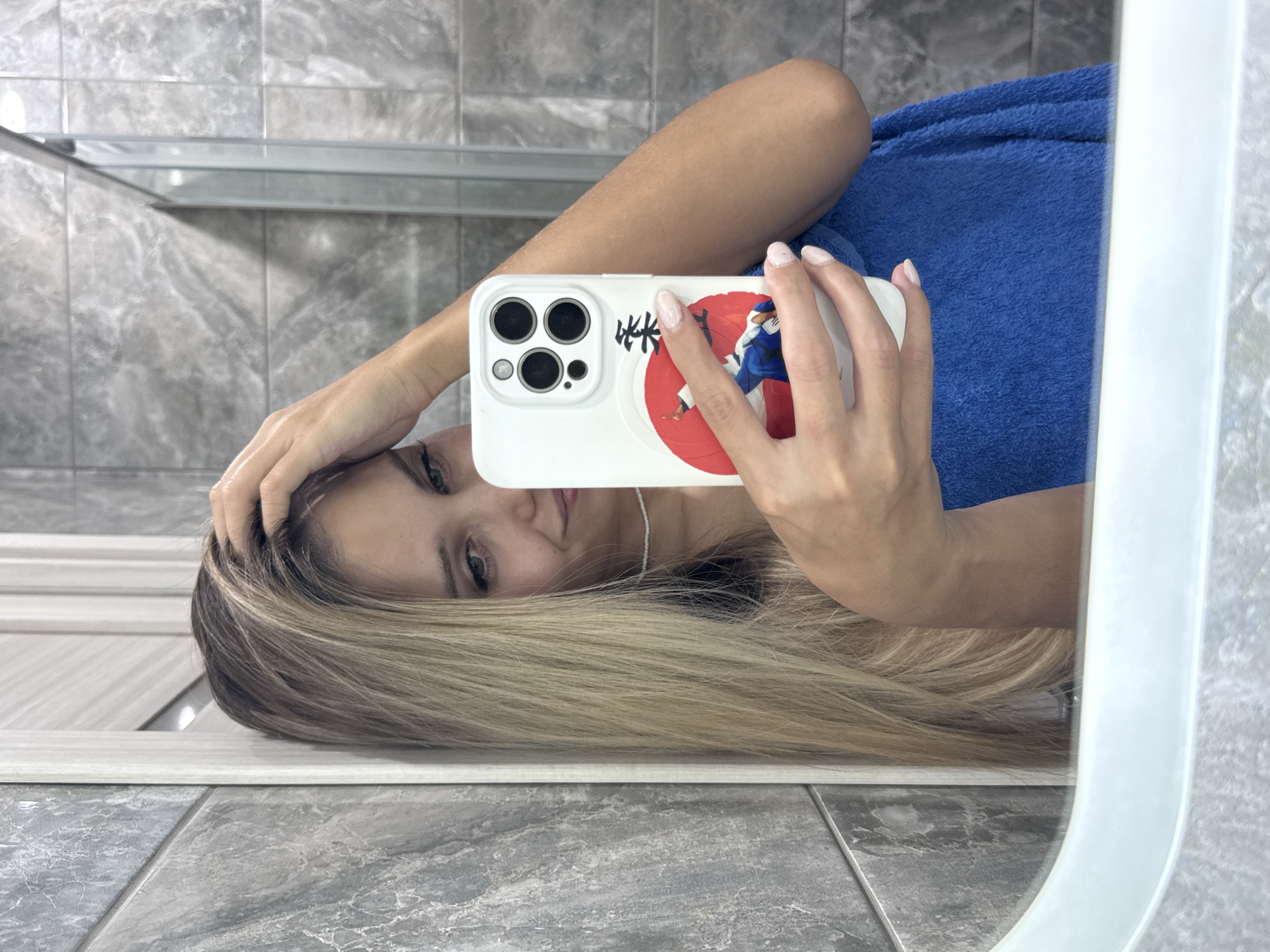 A woman is taking a selfie in a bathroom. The bathroom has gray tiled walls and floors. The woman is holding an iPhone with a white case and a judo sticker. She has blonde hair and is wearing a blue towel. Her left hand is running through her hair.
The location of the image is not provided.