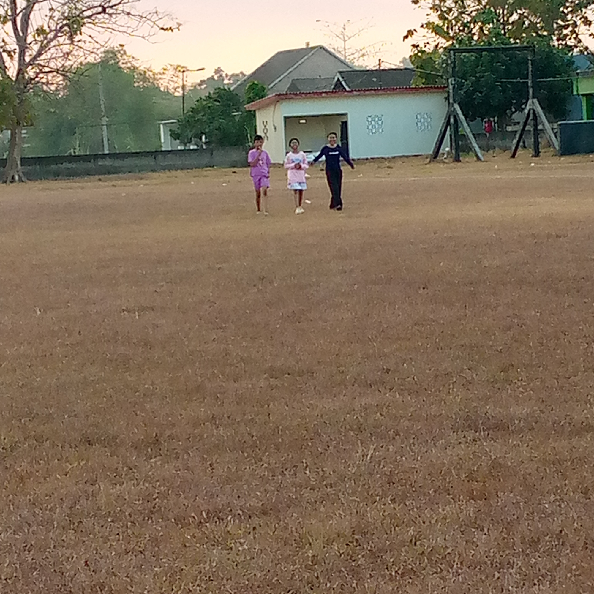 Three individuals are walking on a dry, grassy field in Pacing, Indonesia. The person on the left is wearing a purple outfit. The person in the middle is wearing a light-colored, patterned dress and white shoes. The person on the right is wearing a black outfit. Behind them, a building with a white facade and a peaked roof is visible, as is a basketball hoop structure. The sky is a pale orange, suggesting it is either dawn or dusk.