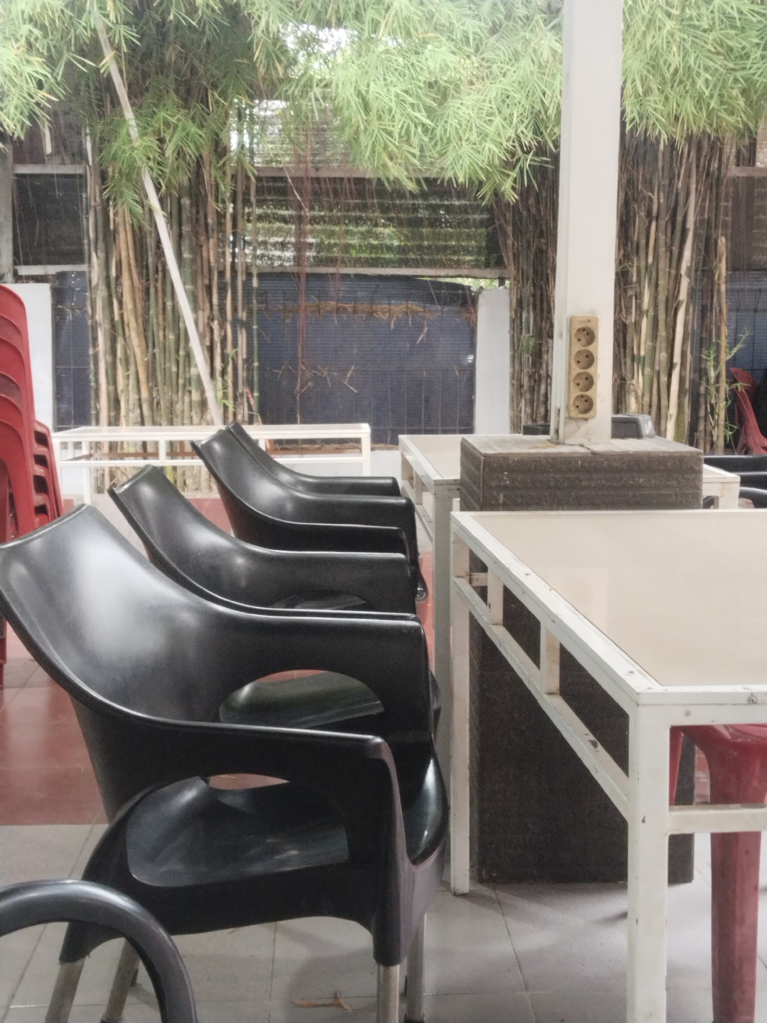 The scene captures an outdoor dining area in Lhokseumawe, Indonesia. Several black plastic chairs with curved backs are positioned around tables with white frames and light-colored tops. The area is semi-enclosed, with bamboo stalks acting as a natural screen along the back. A white column with an electrical outlet is visible. The floor is tiled in a reddish-brown pattern. Several red chairs are stacked in the background.