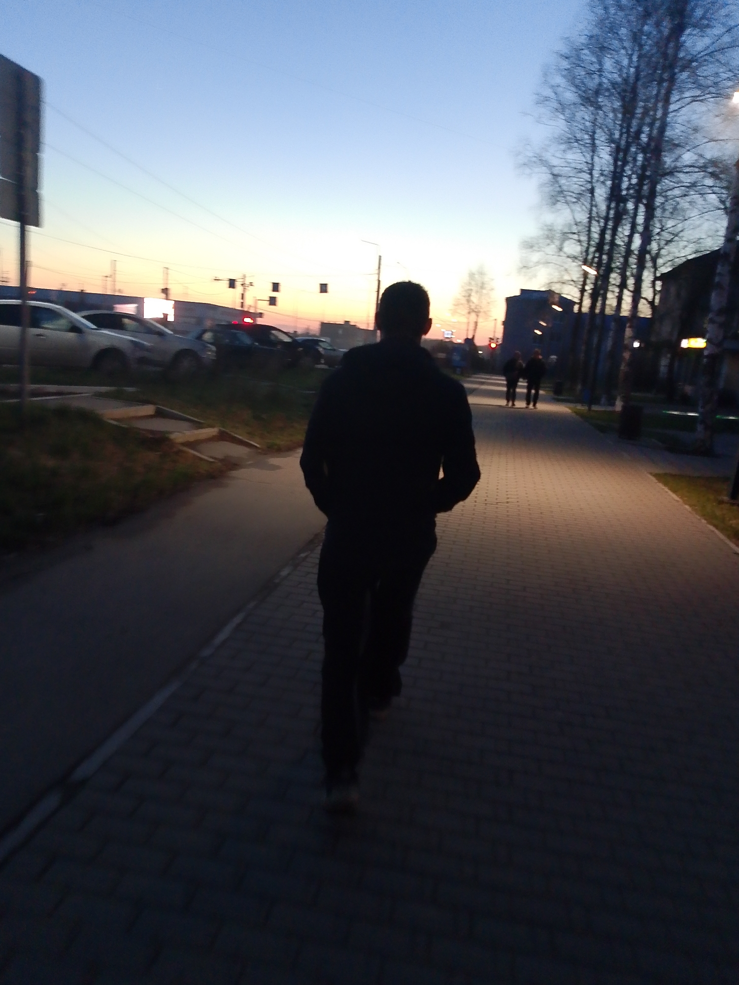 The image depicts an outdoor scene at dusk in Chaikovsky, Russia, viewed from behind a person walking. The primary subject is a male figure, seen from the back in silhouette, wearing a dark hoodie and dark pants, walking on a paved pathway. His figure is sharply contrasted against the soft, fading light of the sky.
The scene is set in what appears to be an urban or suburban environment. The paved pathway, constructed from grey-brown rectangular pavers, extends into the distance. Two other silhouetted figures are visible walking further down the same path. To the left of the pathway, there is a strip of unpaved ground with patchy grass, leading to a road or parking area. Several cars are parked or in motion on this road, including a light-colored sedan in the foreground and darker vehicles further back. A faint red light, possibly from a brake light or a distant signal, is visible among the cars.
The sky, transitioning from a light blue overhead to a warm orange and yellow hue near the horizon, indicates the twilight hours, likely dusk. Bare deciduous trees, characteristic of late autumn or early spring, line the right side of the pathway, with structures and buildings visible behind them. Streetlights are present along the path and in the distance, though their illumination is not strong, complementing the natural fading light. Utility poles and power lines are visible against the sky in the upper left quadrant. No discernable text is visible. The overall mood is calm and transitional as day gives way to night.
