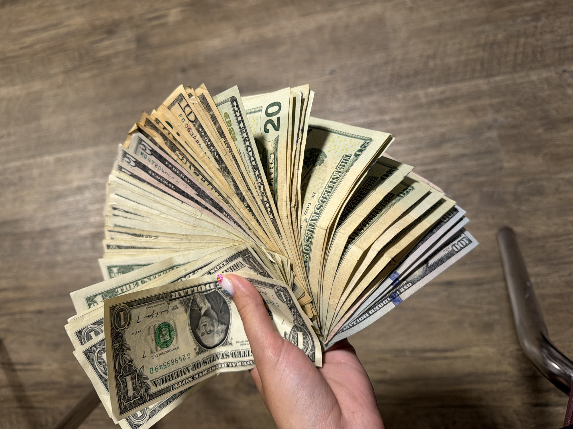 A close-up, eye-level, vertical shot shows a person's left hand fanned out with a large sum of US currency. The bills appear to be a mix of denominations, including what looks like $1, $5, $10, $20, and $100 bills. The hand is holding the bills in a fanned-out manner, with the currency spread across the frame. The background consists of a wooden surface, likely a table or floor, with a visible grain pattern. The lighting appears to be ambient, casting soft shadows. There is no discernible location in Lake Delton, United States of America visible in the image.