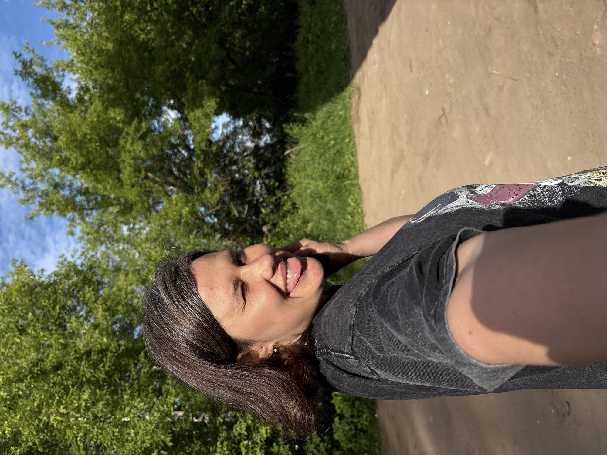 A young woman with dark hair is taking a selfie outdoors in Chaikovsky, Russia. She is wearing a dark t-shirt and is leaning back with her eyes closed and tongue sticking out, appearing playful. Her hand is resting on her head. The background shows lush green trees and a bright blue sky with some clouds, indicating it is daytime and likely pleasant weather. A sandy or dirt path runs diagonally through the scene. The image is captured in portrait orientation.