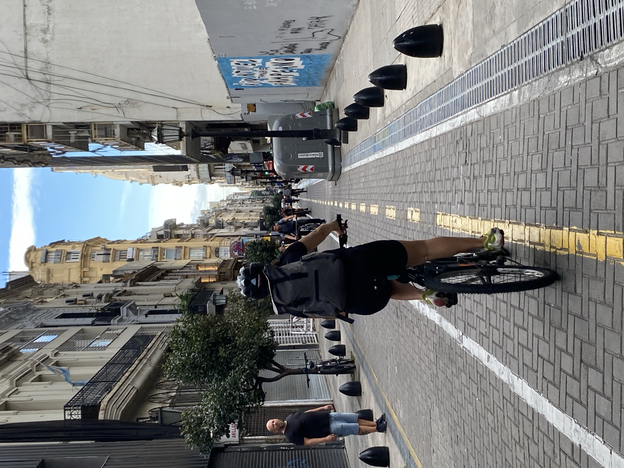 The image shows a street scene in Buenos Aires, Argentina. The perspective is along a long street flanked by tall buildings with ornate facades. The sky is visible in the distance, indicating an opening in the urban canyon.
A cyclist wearing a helmet, a backpack, and dark clothing rides towards the viewer on a bicycle lane marked by yellow lines on a brick-paved street. On the right side of the bicycle lane, there are black bollards lined up along a drainage gutter.
On the left side of the bicycle lane, there's a bald man in a dark t-shirt and blue shorts standing near a tree and parked bicycles. The buildings on the left are older and have balconies.
Further down the street, there are people walking and other cyclists. There's also a dumpster with graffiti on the side. A blue-painted wall with more graffiti is visible in the background.