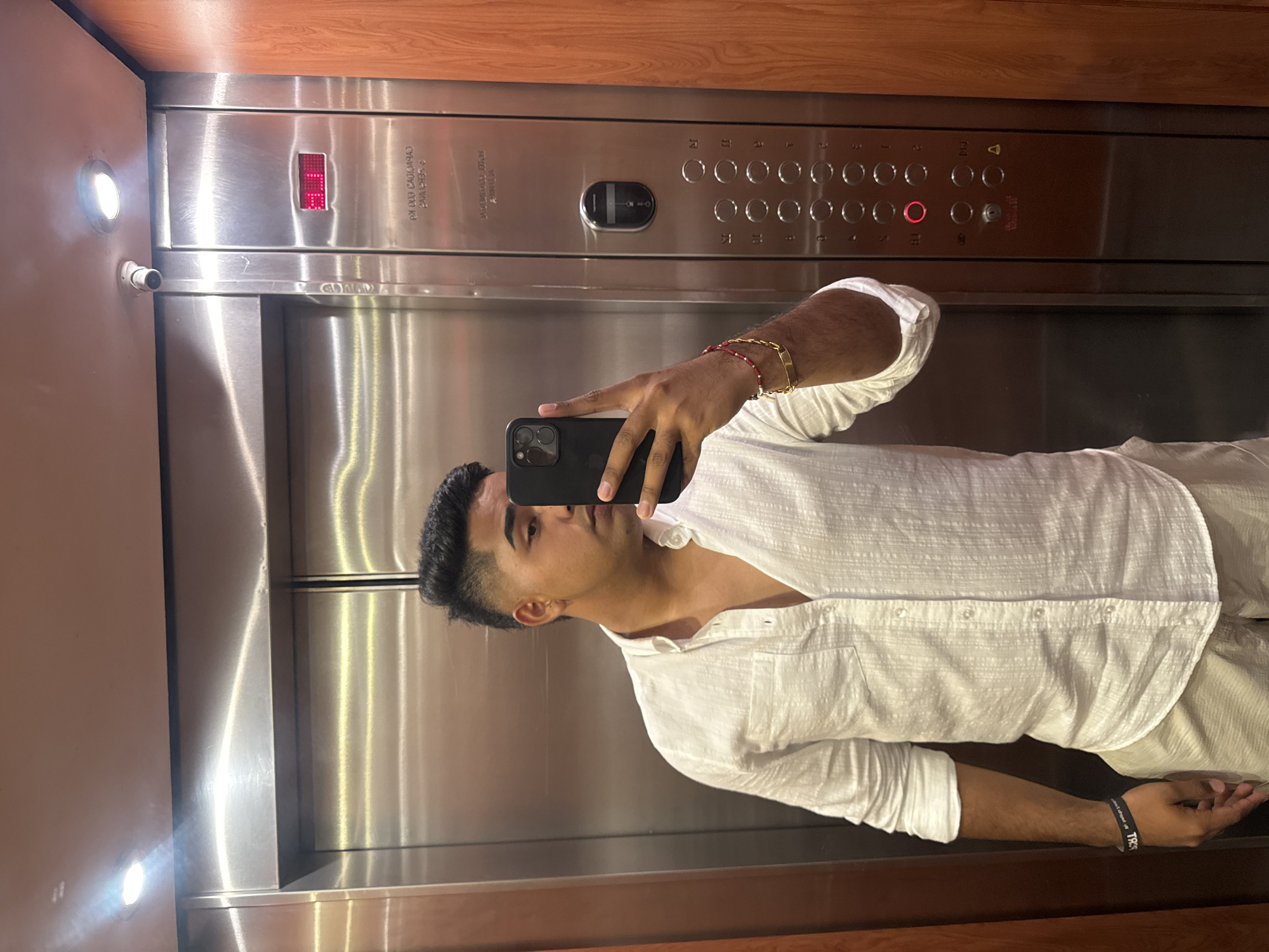 A man is taking a selfie in an elevator. The elevator has a metal interior with wooden paneling on the top and bottom. The control panel shows buttons for floors 1-12, with the "8" button illuminated in red. The man is wearing a white shirt and is holding a black smartphone in front of his face. The location is not evident from the image.