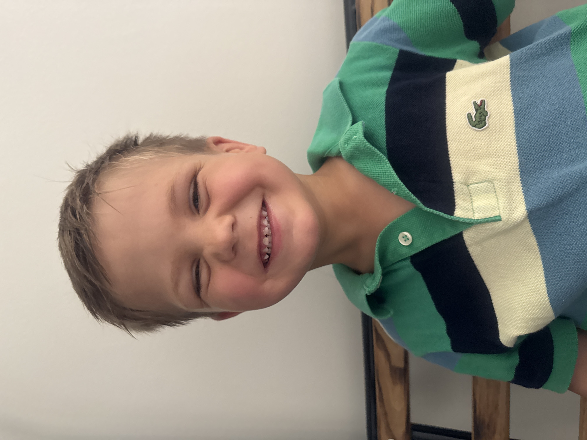 A young boy is smiling with his eyes squinted shut, revealing gaps in his front teeth. He is wearing a striped polo shirt with green, navy blue, and light blue bands, and a small green crocodile logo on the left side of his chest. The boy is positioned in the foreground of the image, with a plain, light-colored wall behind him. The overall scene appears to be indoors. The image does not provide any information about the city of Les Rousses, France, or its specific location within the city.
