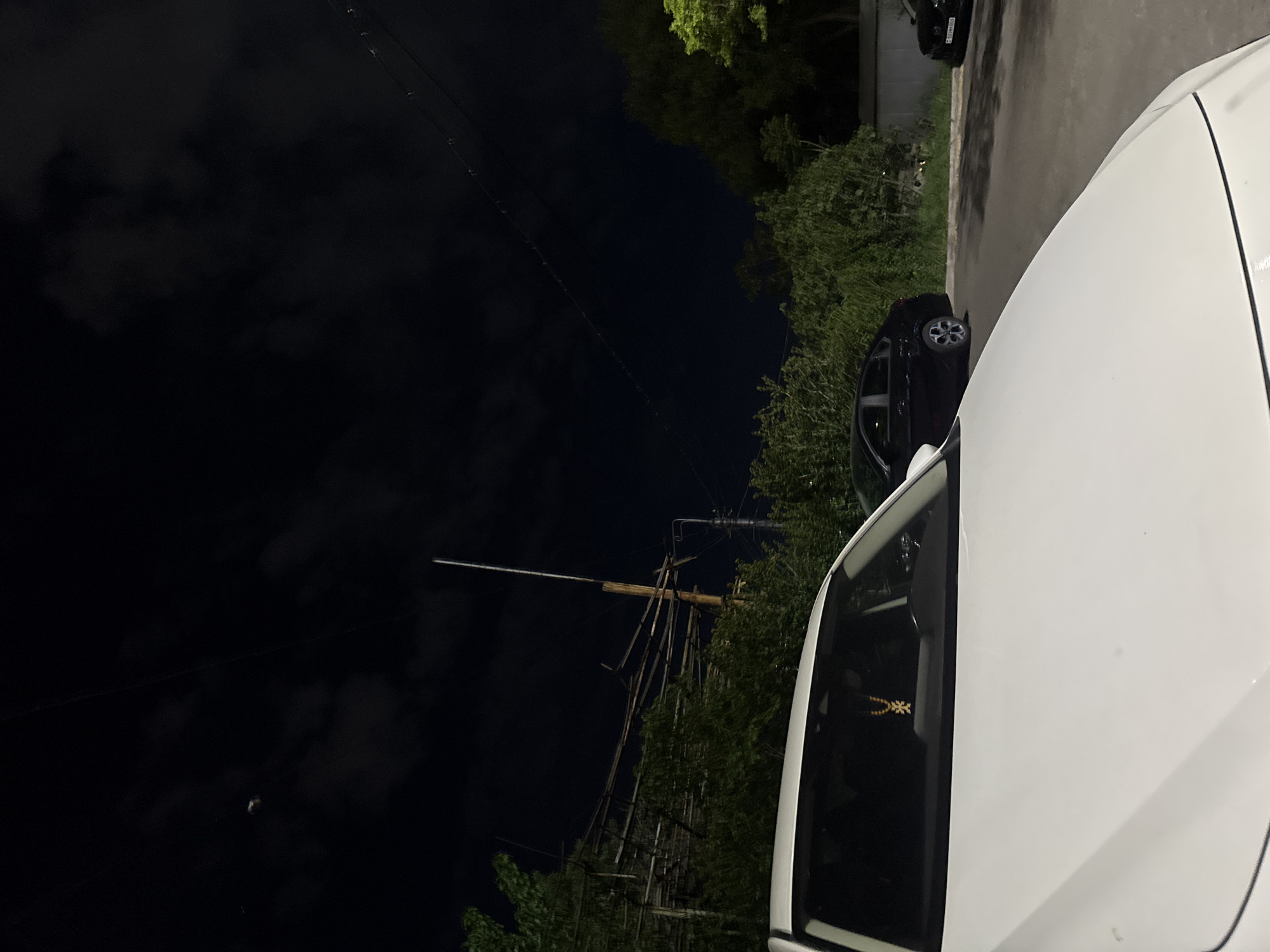 The image is a nighttime view taken at street level. In the foreground, a significant portion of a white car occupies the right side of the frame. Parked adjacent to the white car is a black car. Further into the background, other parked vehicles can be seen. The road is paved and appears wet, likely from recent rain. On the opposite side of the road, there is greenery with what seems to be a crude bamboo construction or support structure visible against a dark sky. Overhead, power lines run across the image. Based on the general appearance and information available, it is plausible that the location may be Yerevan, Armenia, though this is not certain without further visual cues or information.