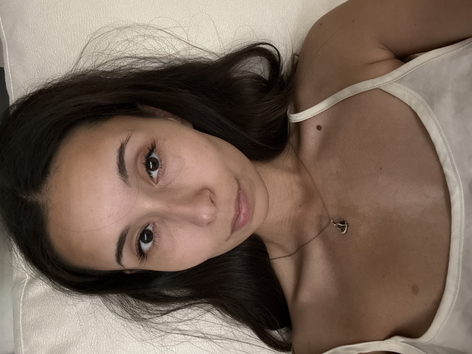 A young woman with dark hair and brown eyes is lying down, looking directly at the camera. She is wearing a white tank top and a delicate necklace with a heart-shaped pendant. The background is a textured, light-colored surface.