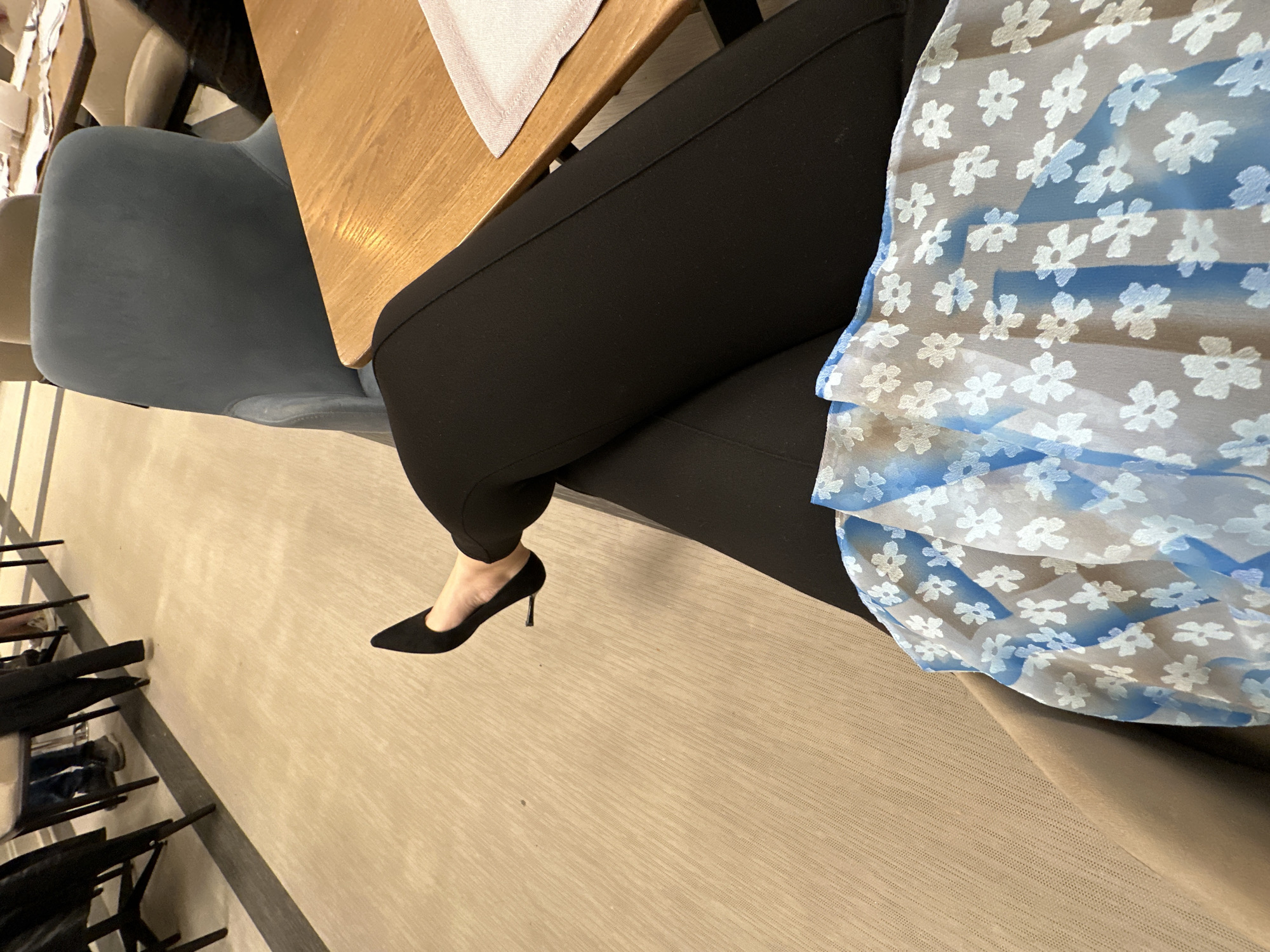 A vertically oriented, high-angle shot depicts a seated individual, likely a woman, at a wooden table in Kazanlak, Bulgaria. She is dressed in black pants and a blue and white floral print top. Her footwear consists of a single visible black stiletto heel. The person is seated in a light blue upholstered chair at the table. The floor beneath her is beige. Additional chairs and tables are visible along the left edge of the image.