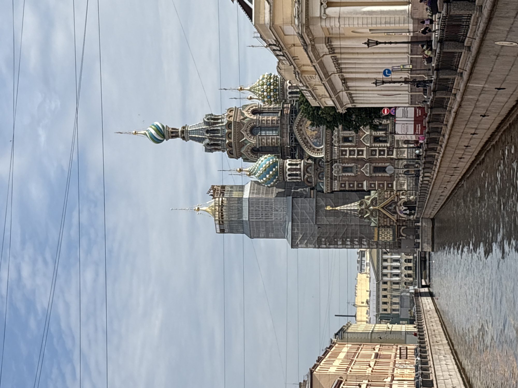 The image shows a canal in Saint Petersburg, Russia, with the Church of the Savior on Spilled Blood visible in the background. The church is a prominent landmark with its colorful onion domes and intricate architecture. The canal is lined with stone embankments and surrounded by buildings that reflect the city's historical architecture. Several people are visible walking along the canal's edge. The sky is cloudy, and the water in the canal is calm, reflecting the surrounding structures. Some of the scaffolding on the Church of the Savior on Spilled Blood suggests ongoing renovations or maintenance. The scene depicts a typical day in the historic city center.