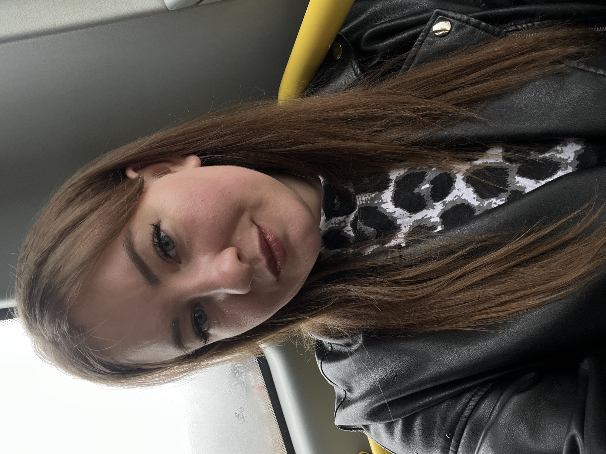 A woman with long, brown hair is seated in a vehicle, likely a bus or train, as evidenced by a yellow handrail visible above her. She is wearing a black leather jacket over a garment with a black and white patterned design. The woman is looking towards the camera with a slight smile. The image was taken in the city of Chaikovsky, Russia. A window is visible in the lower left corner.