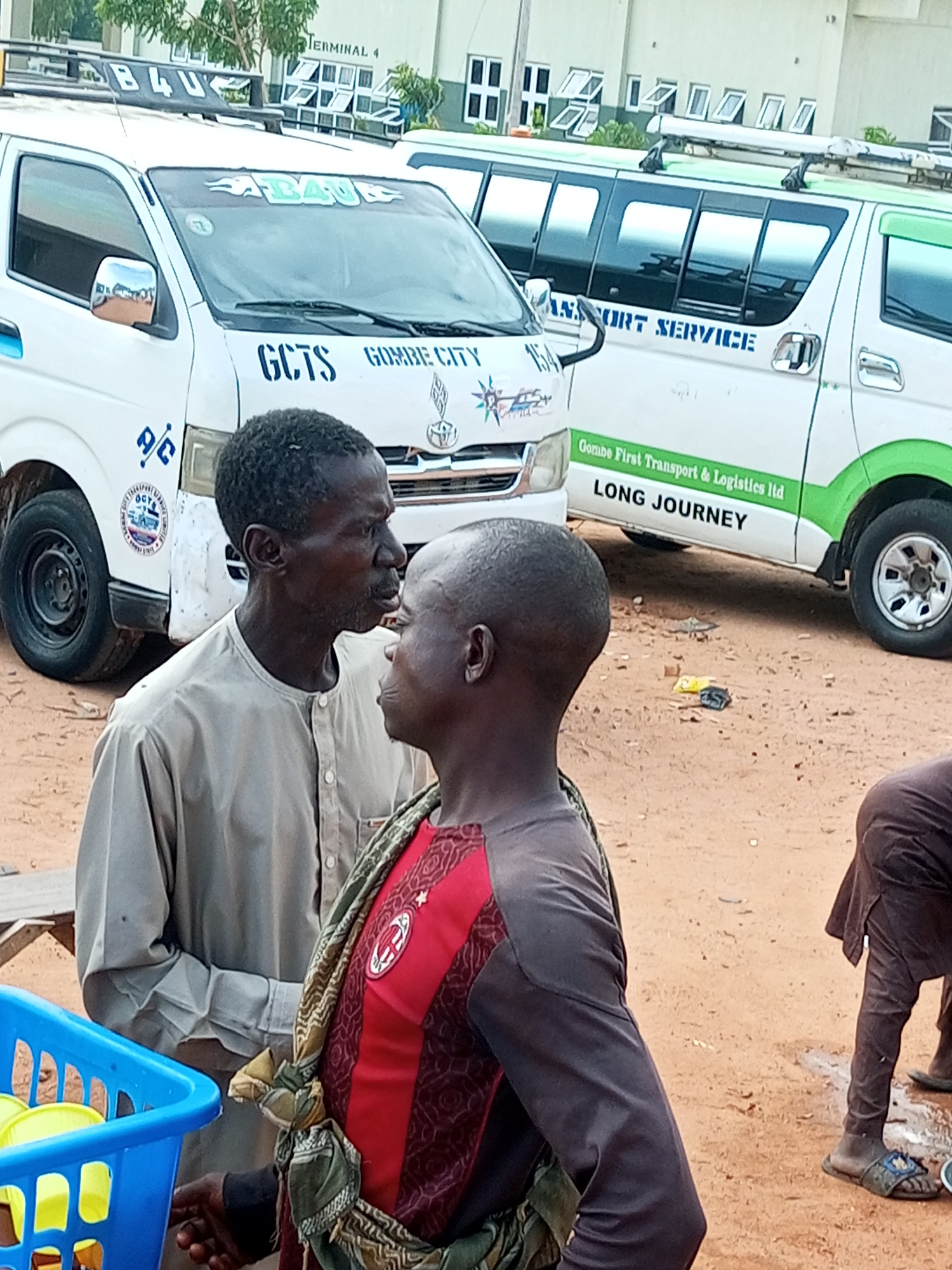 Two men stand in front of a white and green bus with "Gombe First Transport & Logistics ltd LONG JOURNEY" written on it. The man on the left wears a light brown, long-sleeved shirt. The man on the right wears a red and black striped shirt and a patterned scarf. A blue plastic basket filled with yellow objects is in the foreground on the left. The ground is sandy and there is some litter scattered around. In the background, another white bus with "B4U" written on it is visible, along with a building labeled "TERMINAL 4".