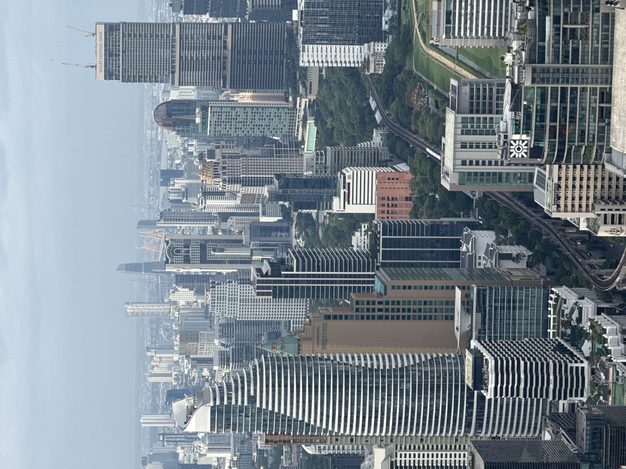 This is an aerial view of a dense urban landscape, likely taken in Bangkok, Thailand. The scene is dominated by skyscrapers and high-rise buildings of varying designs and heights, tightly packed together. The architectural styles range from modern glass facades to buildings with distinct patterns and textures. Amidst the buildings, there is a green area suggesting a park or gardens with elevated train tracks weaving through the urban environment. The sky is hazy, contributing to the overall impression of a sprawling metropolis.