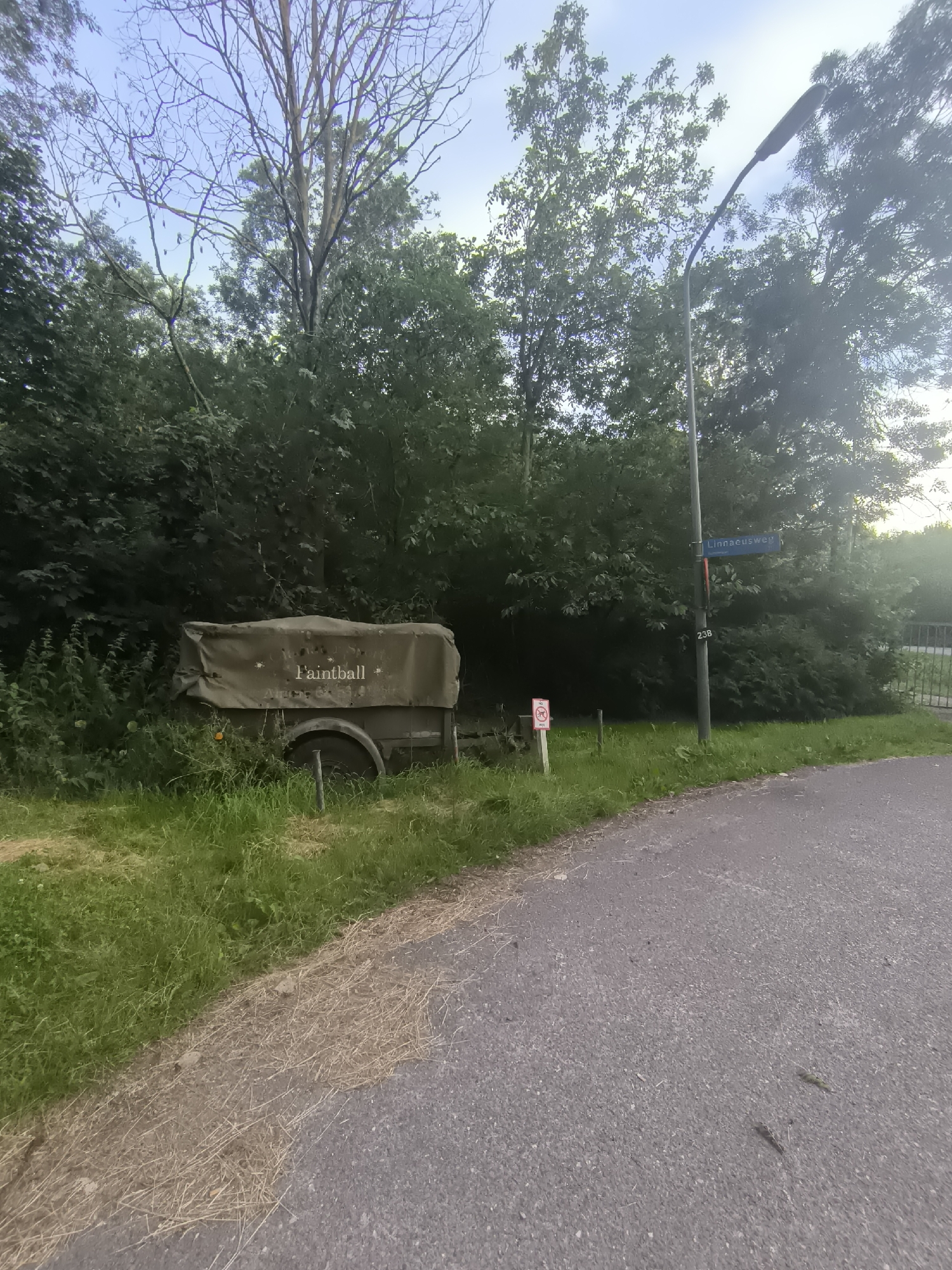 In Almere, Netherlands, along a road adjacent to dense greenery, a weathered, tarp-covered trailer with the word "Paintball" visible on its side sits in the grass. A streetlamp stands nearby, accompanied by a blue street sign indicating "Linnaeushweg" and a pole with the number "238." To the left of the trailer, a small white sign with a red circle and a diagonal line through a silhouette of a person is visible. The scene appears to be a roadside setting in a suburban or park-like area.