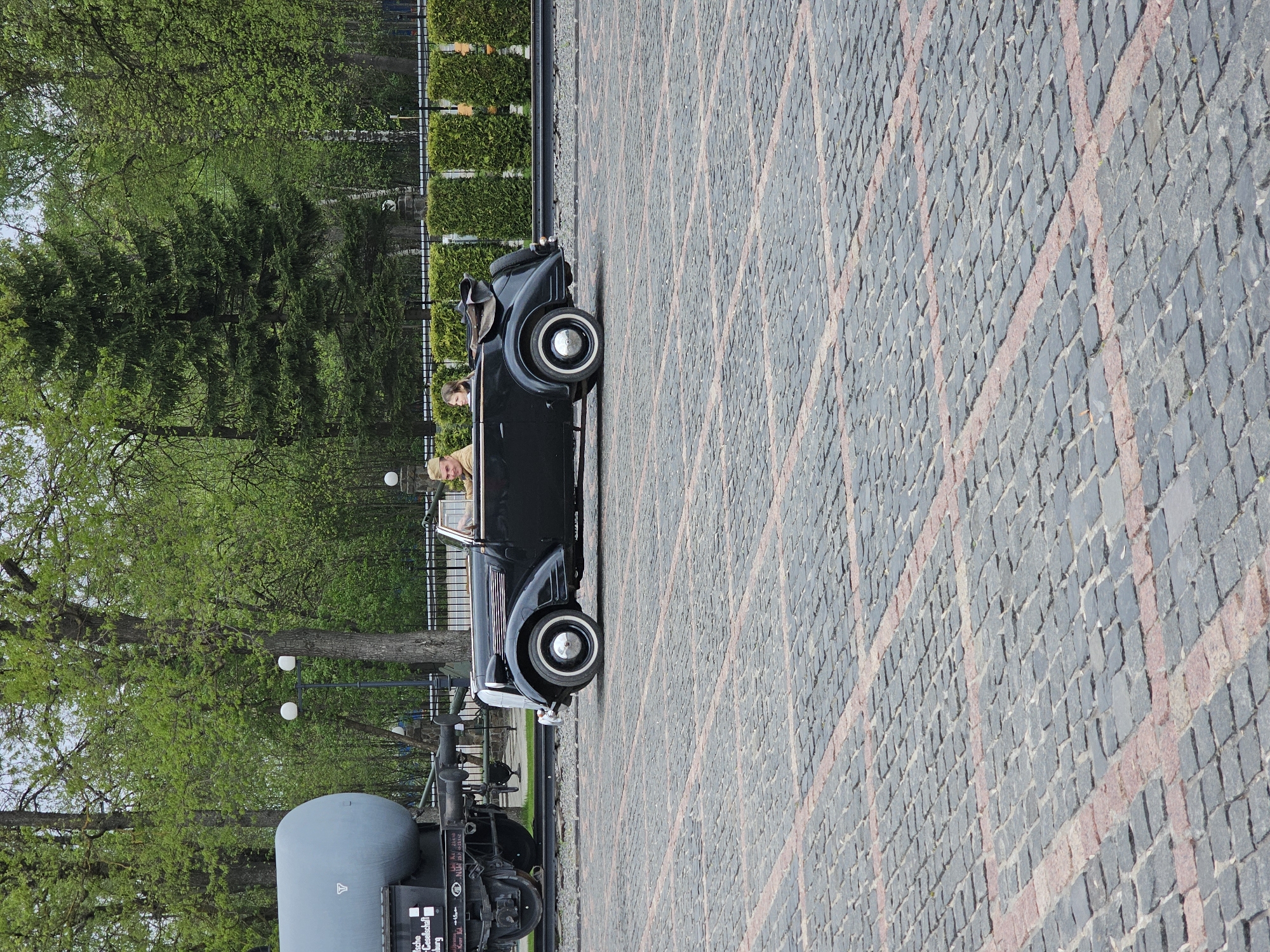 The image depicts a vintage black convertible car with two occupants, parked on a cobblestone street with red brick accents. The car is situated next to a static display of vintage train equipment, including a gray tank car. Lush green trees line the background, suggesting a park or garden setting. The scene appears to be located outdoors in an urban or historical area, given the style of the car, the train equipment, and the landscaping. Based on additional information the location could be the city of Arkhangelskoye, Russia.