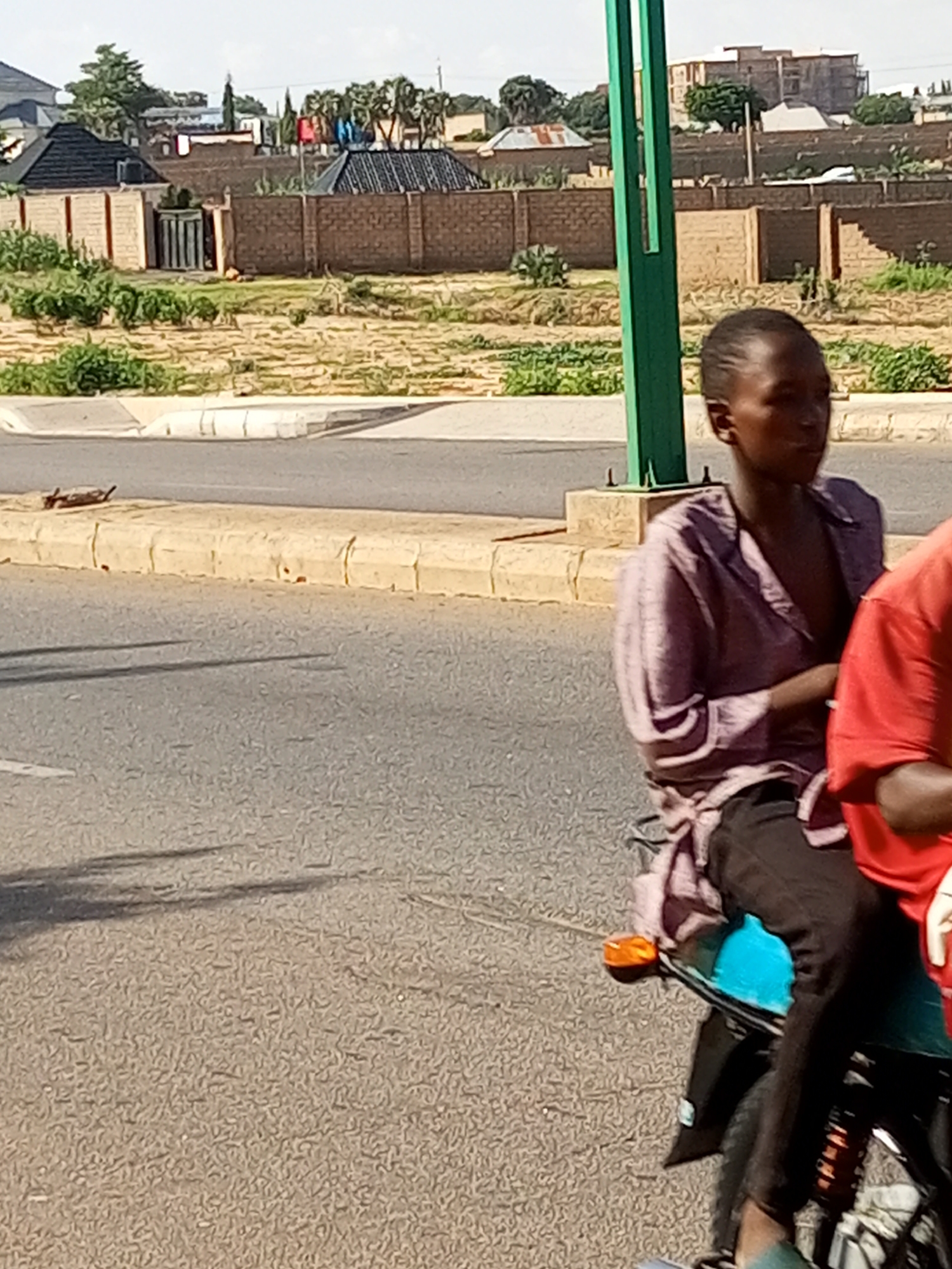 In the city of Gombe, Nigeria, a light-skinned individual wearing a lavender-colored button-down shirt and dark pants is seated on the back of a motorcycle. The person's left arm is bent, resting on their lap. To their right, a portion of another person wearing a red shirt is visible. The motorcycle has a bright turquoise fender. The scene is captured during daylight on a paved road, with a raised concrete median separating the roadway from a dirt area lined with a brick wall. In the background, there are buildings, trees, and a green pole.