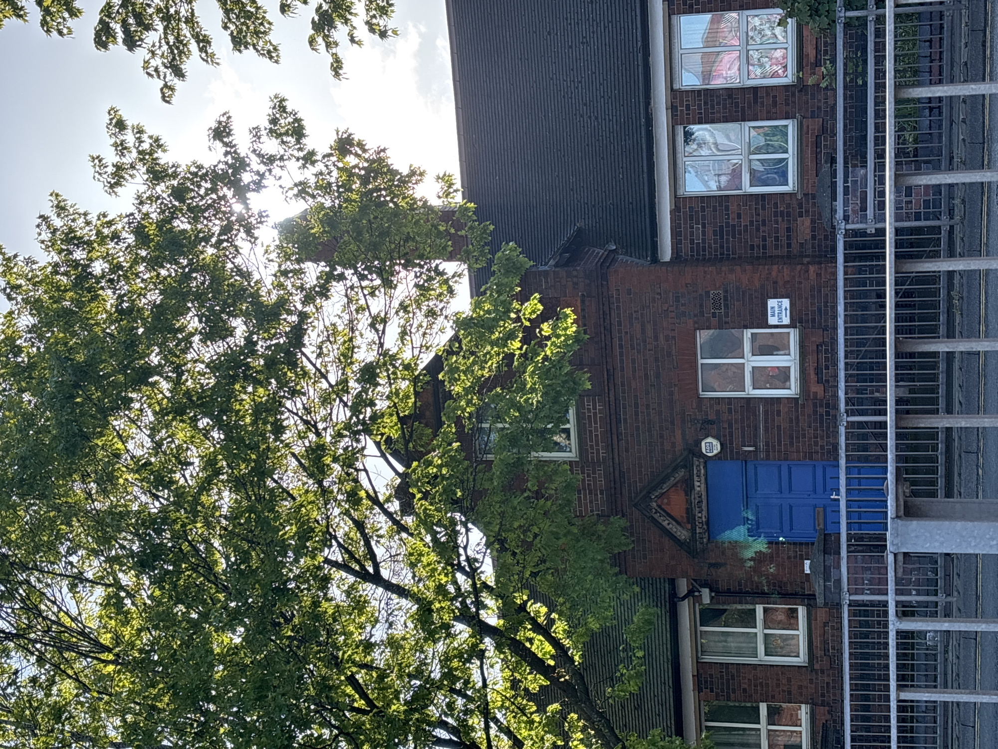 A daytime view of a building, likely a school in Newcastle-under-Lyme, United Kingdom, partially obscured by a tree. The building is constructed of red brick with a dark, corrugated roof. There are multiple windows, some with patterned coverings or artwork inside. One window displays what appears to be stained glass. There is a blue door with a triangular architectural feature above it. A fire escape ladder is visible on the right side of the building. A sign, partially obscured, is visible near one of the windows. The sun is shining through the tree branches.