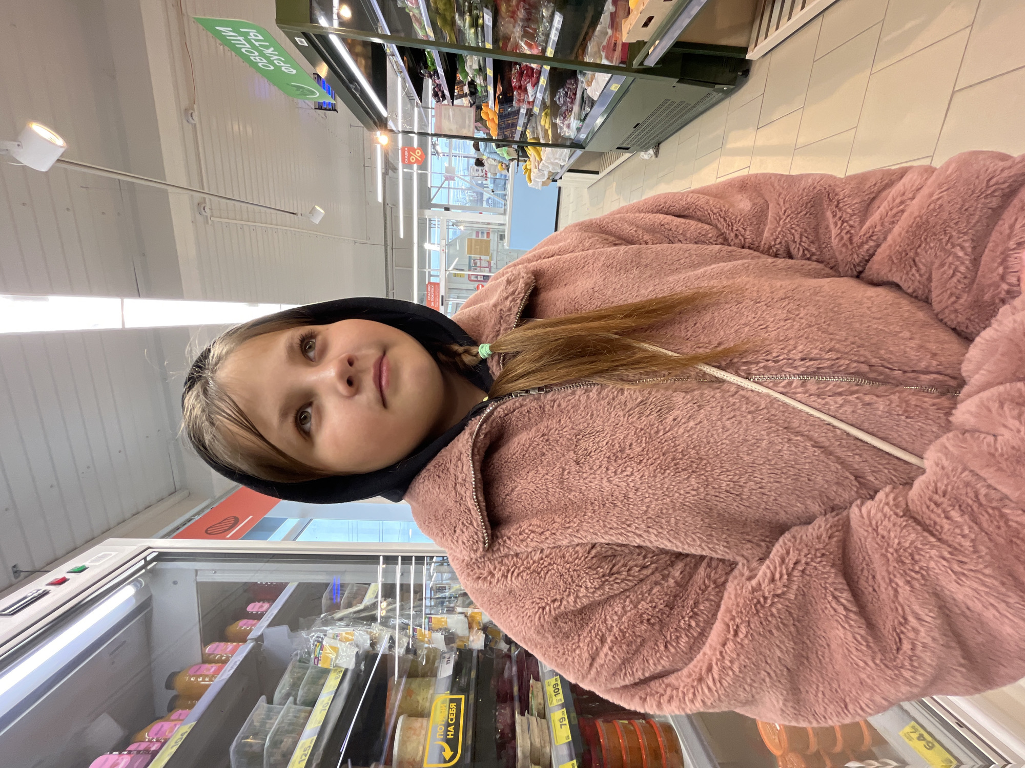 A young girl wearing a pink fleece jacket with a black hood is in a grocery store in Novopokrovskaya, Russia. She is standing in front of a refrigerated display case containing various food items. The store has fluorescent lighting and a white tile floor. Signs in Russian are visible. The display case contains food items of different colors. Other store aisles and shelving are visible in the background.