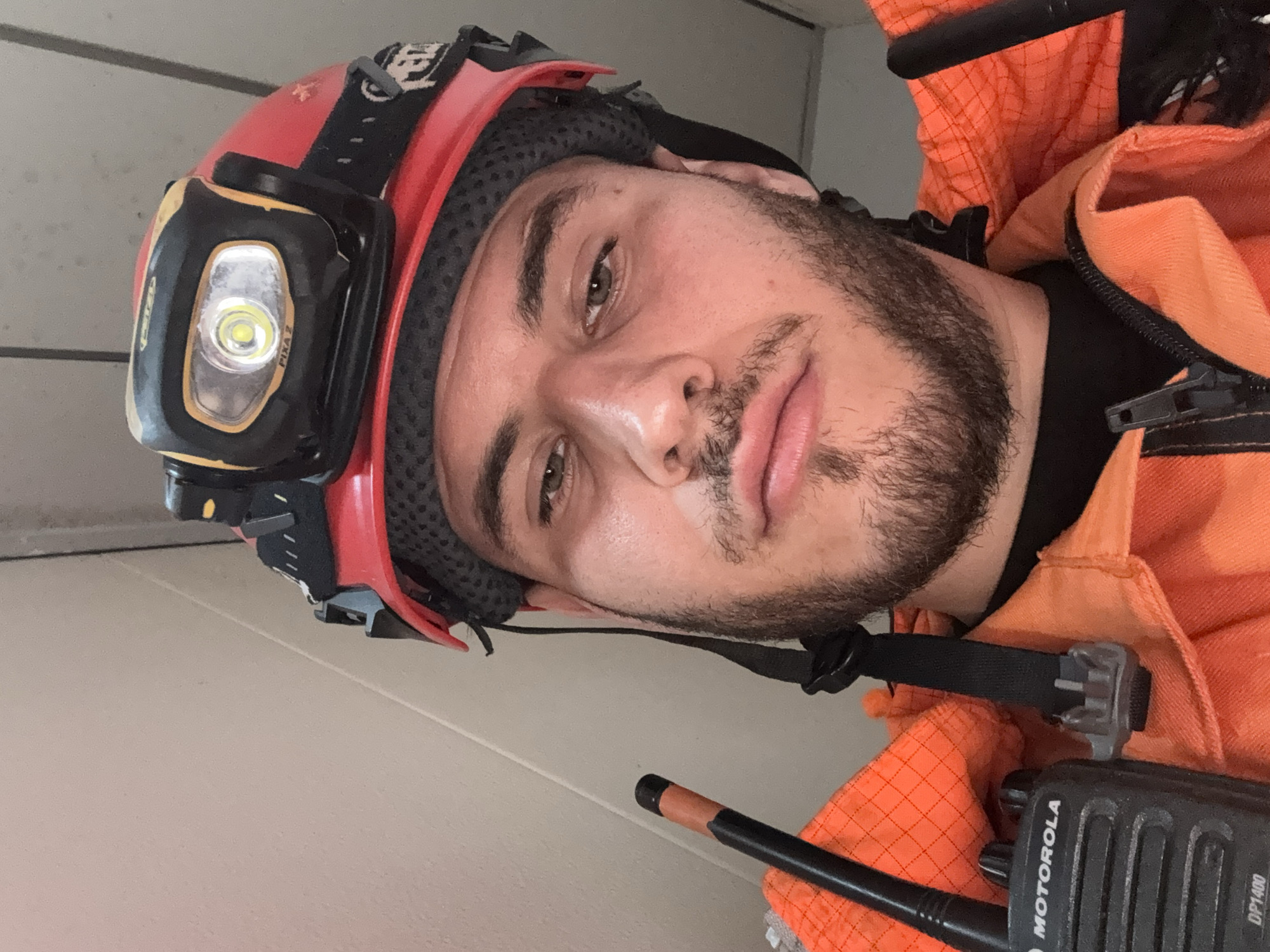 A man wearing a red safety helmet with a headlamp and an orange jacket is captured in a close-up selfie. The man has a beard and fair skin. He is also wearing what appears to be a Motorola DP1400 two-way radio. The background appears to be a plain, light-colored wall. The location of this image is unknown and cannot be determined from the image.