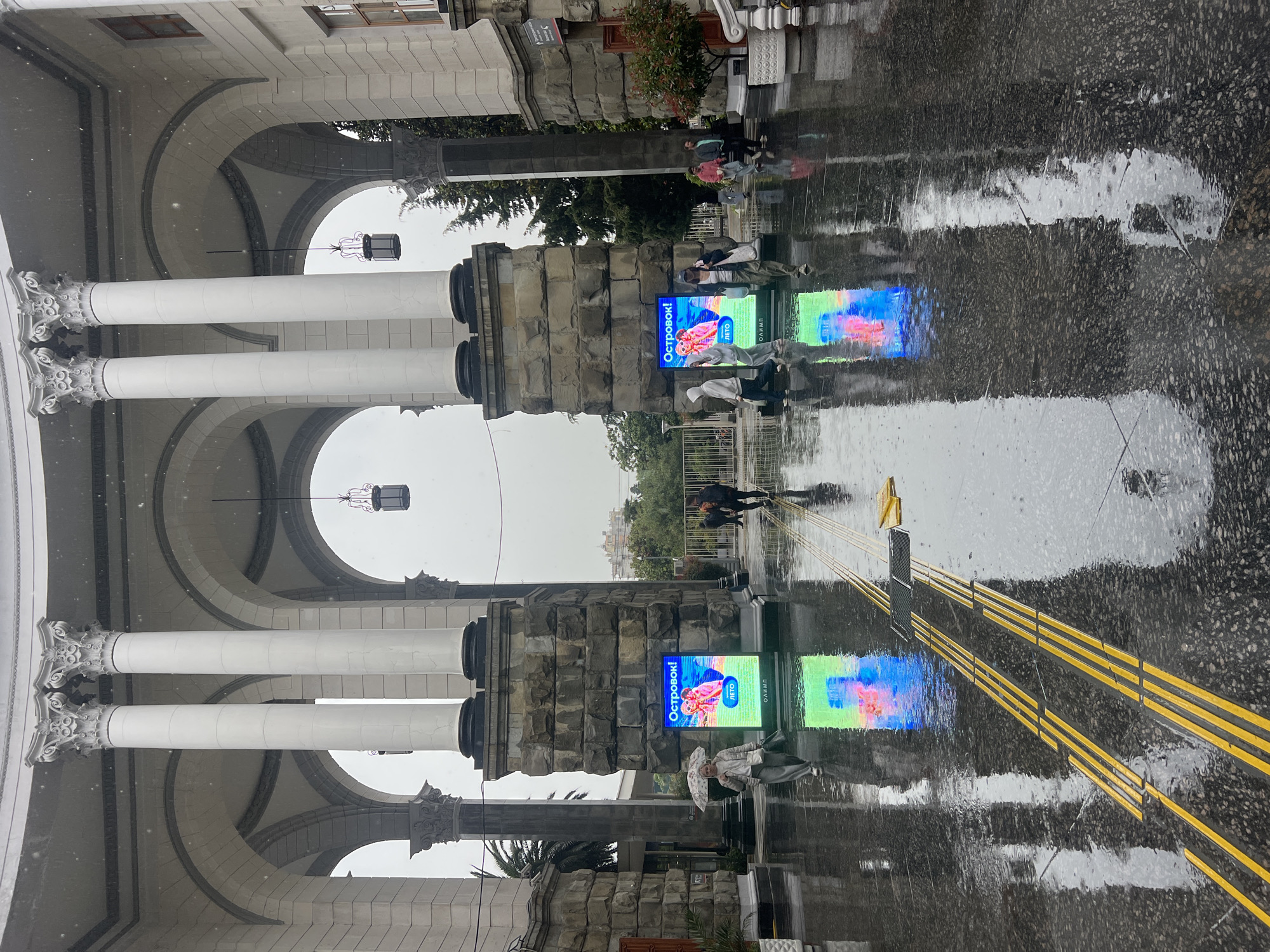 The image shows a reflection of a building on a wet surface in Sochi, Russia. The building features a colonnade with white pillars and arches. There is a person walking in the background. An advertising screen is displayed on a post next to the colonnade. The ground has yellow lines.