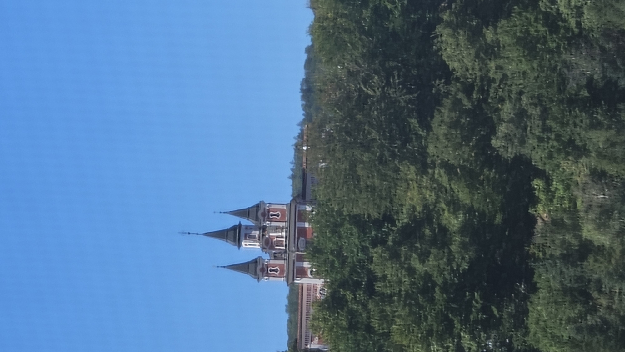A red and white church with three conical spires stands atop a hill covered in lush green trees. The sky is a clear, bright blue. The church has a prominent central tower with a smaller spire, flanked by two slightly shorter towers with darker spires. The building features red walls with white trim and decorative elements. A long, low section of the building extends to the right, also topped with a red tiled roof. The foreground is dominated by the dense foliage of trees, obscuring the base of the hill. The image appears to be taken from a distance, looking up towards the church. This structure is the Orthodox Church of the Holy Spirit in Prešov, Slovakia.