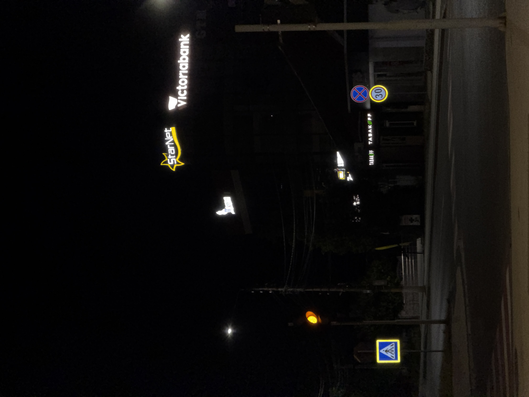 The image captures a nighttime street scene in Strășeni, Moldova. A yellow pedestrian crossing sign is visible on the left, illuminated by a traffic light showing a yellow signal. In the background, the illuminated signs for "StarNet" and "Victoriabank" are prominent. Further along the street, signs for "TABAKOFF" are also lit. Two circular traffic signs are present: one indicating no stopping, and another displaying a speed limit of 30. The street is asphalted, and a crosswalk is faintly visible in the foreground. The overall environment is dark, suggesting nighttime, with artificial lighting from signs and streetlights.