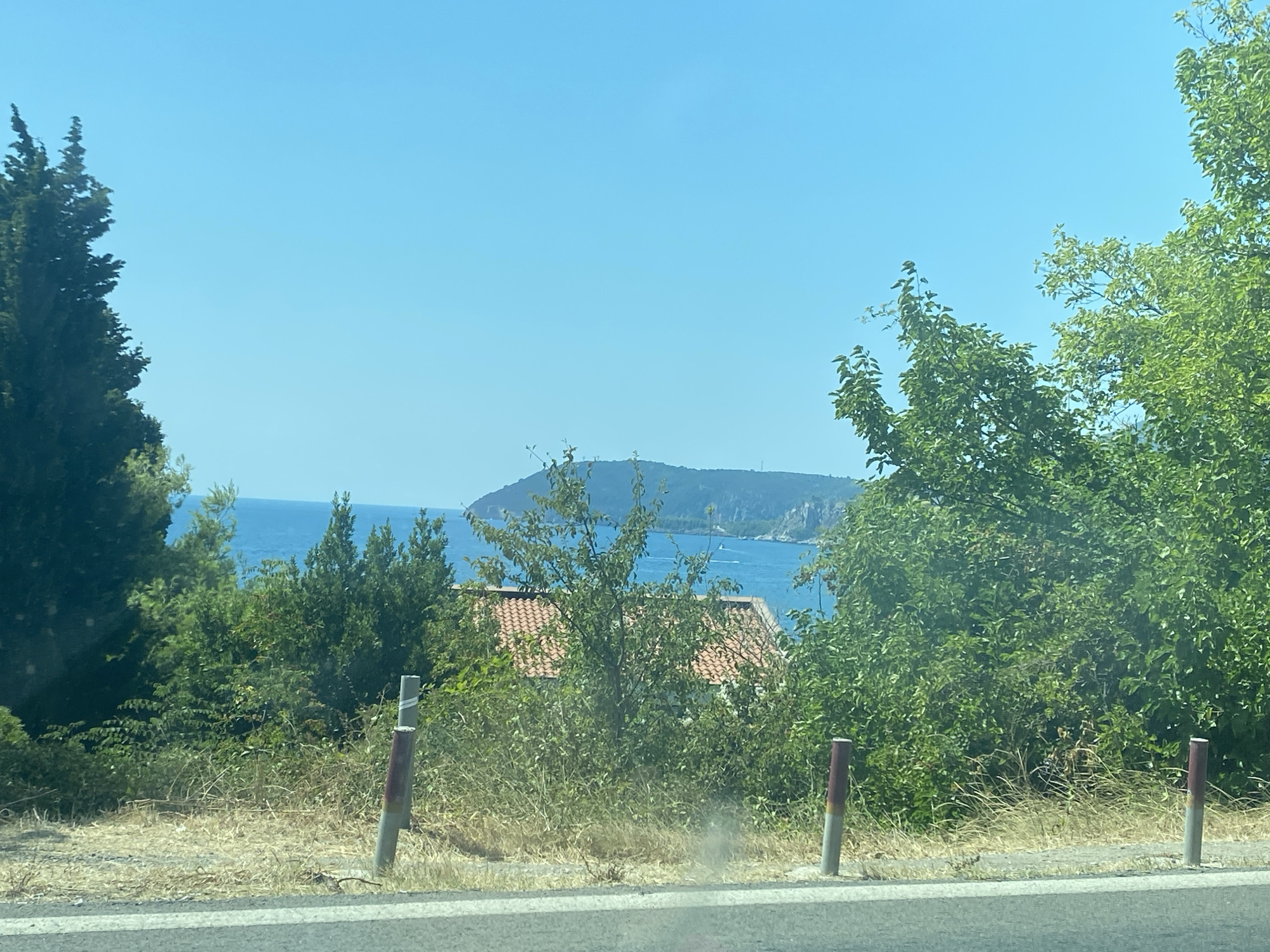 The image captures a scenic view from a roadside vantage point, likely in the city of Sutomore, Montenegro. The foreground features a dry, grassy verge with two cylindrical posts, one on the left with a red and white striped band and another on the right with a single red band. A paved road surface is visible at the very bottom of the frame.
Beyond the verge, a dense green shrubbery and trees dominate the mid-ground. Peeking through the foliage, the orange-tiled roof of a building is visible, suggesting a residential area.
In the background, a body of clear blue water, likely the Adriatic Sea, stretches towards a distant, rocky coastline or peninsula. The water is calm, with faint white streaks indicating distant boat wakes. The sky is a clear, vibrant blue, typical of a sunny day. The overall impression is one of a tranquil coastal landscape.