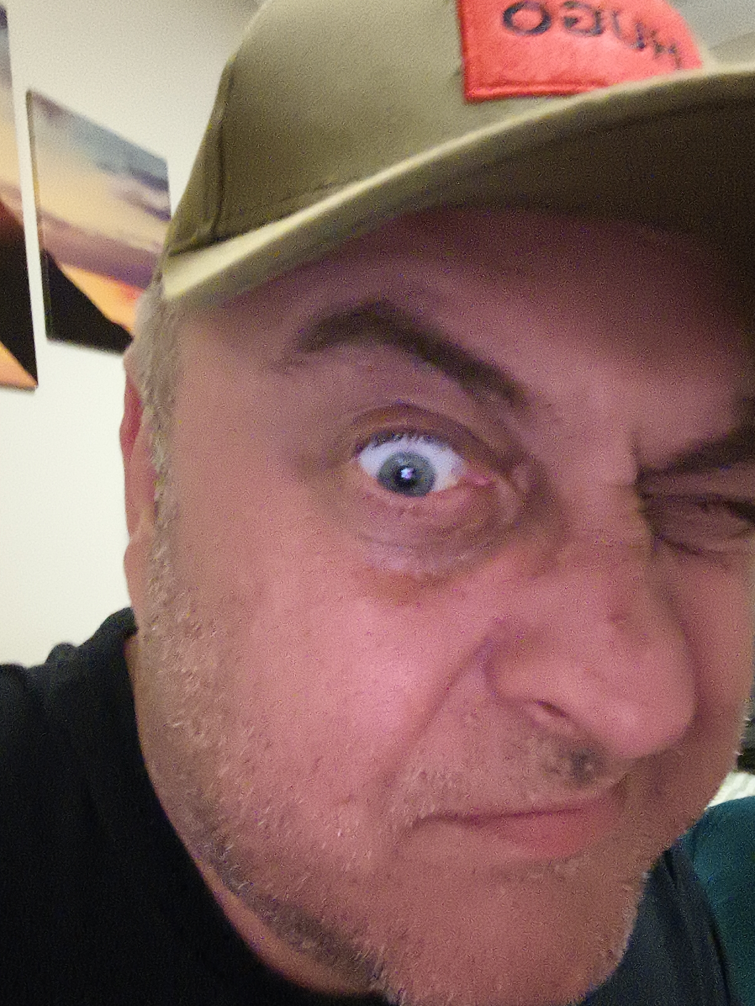 A close-up, eye-level portrait features a middle-aged man with light stubble on his face, making a playful expression. His left eye (viewer's left) is wide open and blue, while his right eye is winked shut, conveying a humorous and quirky mood. He is wearing a dark t-shirt and an olive green baseball cap. A red patch on the front of the cap displays inverted black text, which reads "HUGO" when viewed correctly.
The background is softly blurred, showing a light-colored wall with indistinct, framed artwork or photographs on the left side, exhibiting abstract shapes and colors including purple, orange, and blue tones. The lighting is bright and frontal, illuminating his face evenly and suggesting an indoor setting. The image captures a candid or staged moment of lightheartedness, likely a selfie. No specific location cues beyond an indoor setting are visible within the frame, but the context indicates Huntingdonshire, United Kingdom.