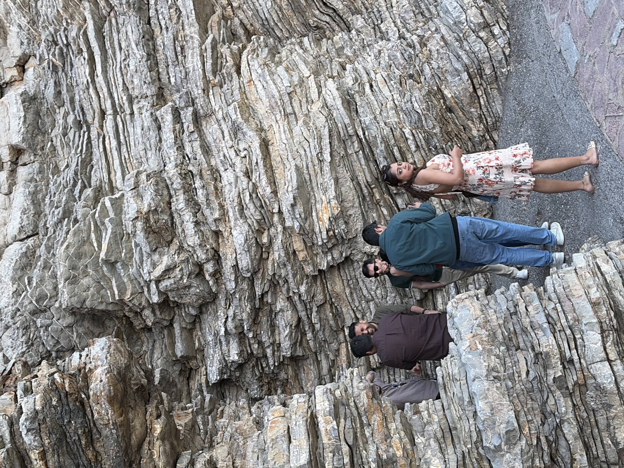 The image captures a group of four people posing in front of a geological formation in Budva, Montenegro. A woman in a floral dress lies down, while three men stand behind her. The location is characterized by layered rock formations with vertical stratification, suggesting a coastal or mountainous setting. The ground transitions to a paved surface towards the top of the image.