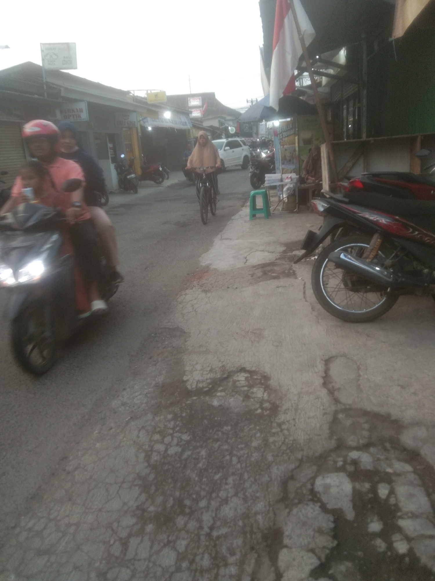 The image depicts a street scene in Subang, Indonesia, with a focus on the road and the surrounding buildings. In the foreground, a motorcycle is visible, with its headlights illuminated, indicating it is either moving or has recently been in use. A person wearing a red helmet is seated on the motorcycle, suggesting they are either the rider or a passenger. To the left of the motorcycle, another person is partially visible, also appearing to be seated.
Further down the street, a person wearing a light brown headscarf and a light-colored outfit is riding a bicycle. They are captured in motion, pedaling along the road. Several other motorcycles are parked or moving in the background, contributing to the impression of a busy urban environment. The buildings lining the street appear to be shops or small businesses, with signage visible, though the text is largely illegible due to the image quality. One sign, partially obscured, shows what appears to be a red and white flag. The road surface is uneven and shows signs of wear and tear, with patches of darker discoloration suggesting moisture or stains. The overall lighting suggests it might be late afternoon or early evening.
