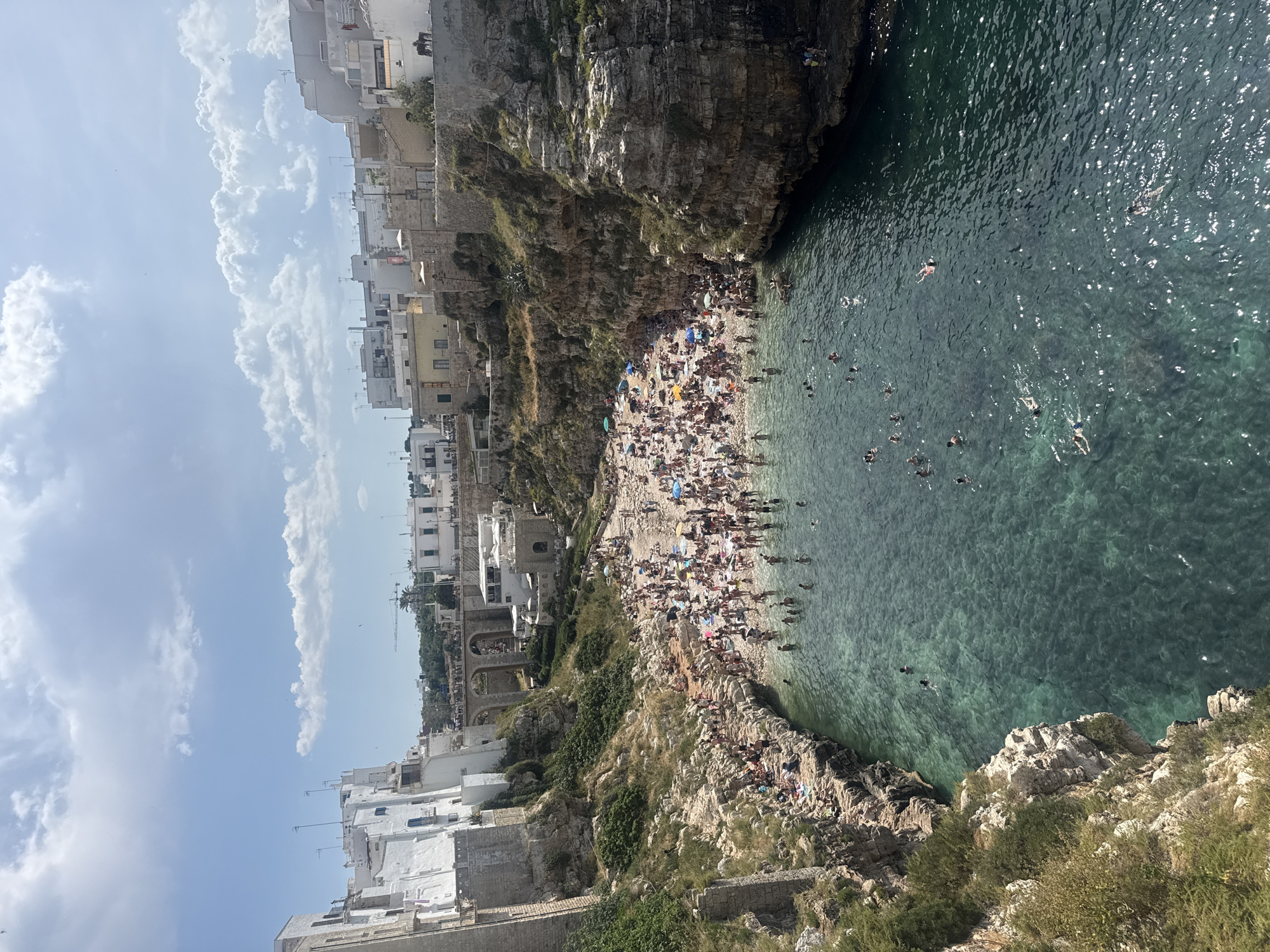 The image depicts a beach in the city of Polignano a Mare, Italy. The beach, located in a cove surrounded by cliffs, is densely populated with people, umbrellas, and towels. Many people are swimming in the clear water of the cove. White buildings line the cliffs above the beach. The sky is blue with scattered clouds. The overall scene captures a busy summer day at this location.