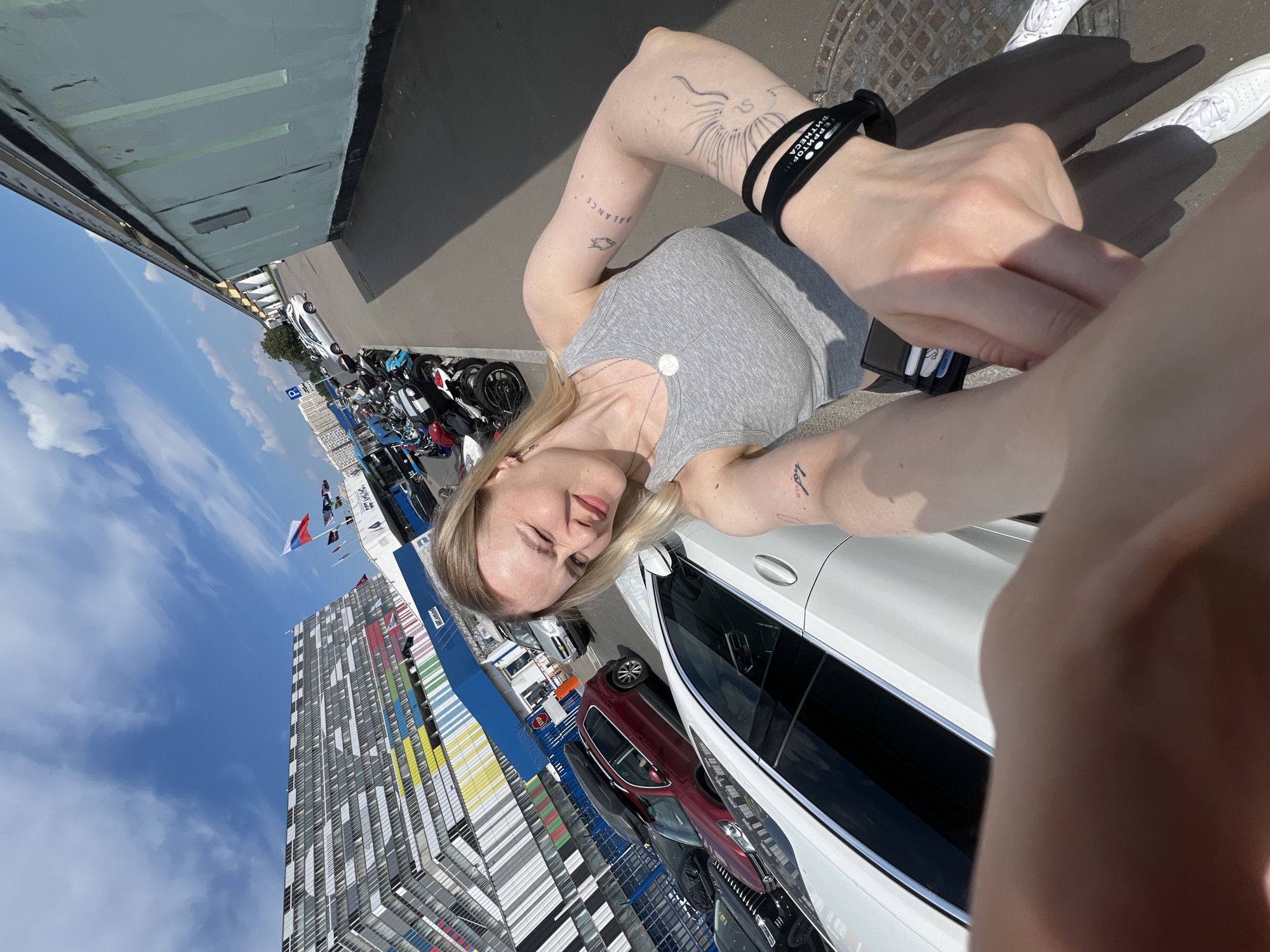 The image shows a young woman with blonde hair, wearing a grey tank top and white sneakers. She has several tattoos on her arms, including one on her left arm that says "balance" and another on her right arm. She is holding a black wallet and a black band with white text on her left wrist.
The woman is standing in what appears to be an outdoor parking lot. Behind her, there are several parked motorcycles and cars, including a white car that takes up a significant portion of the foreground. In the background, there is a tall, modern building with a colorful facade, featuring patterns of white, grey, yellow, and blue. Various flags are visible on poles in front of the building. The sky is blue with scattered white clouds, suggesting it is daytime and the weather is pleasant.
The overall impression is a candid, possibly selfie-style photo taken on a sunny day in an urban environment. The woman's expression is a slight smile, and she seems relaxed. The context of Khimki, Russia, places the scene in a specific geographical location. No other individuals are clearly visible in the image, though distant figures might be present within the parking lot or near the building. The image captures a moment of casual outdoor activity.