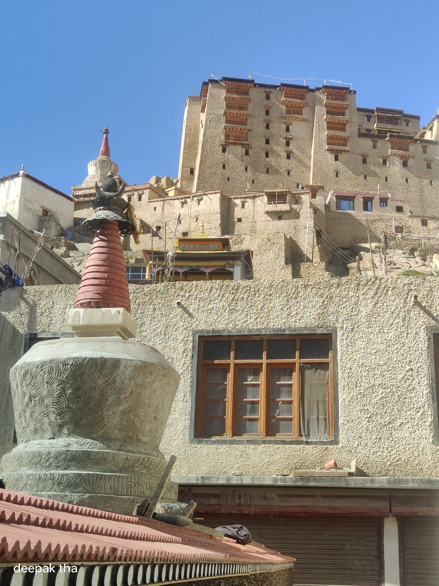 A Buddhist stupa stands in the foreground of the image, against a backdrop of the Leh Palace in Leh, India. The stupa is made of stone and is decorated with intricate carvings. It has a base, a dome, and a spire. The Leh Palace is a tall, multi-story building made of stone. It has a distinctive architectural style, with many windows and balconies. The sky is clear and blue, and the sun is shining. The scene is serene and peaceful, conveying a sense of tranquility and spirituality.