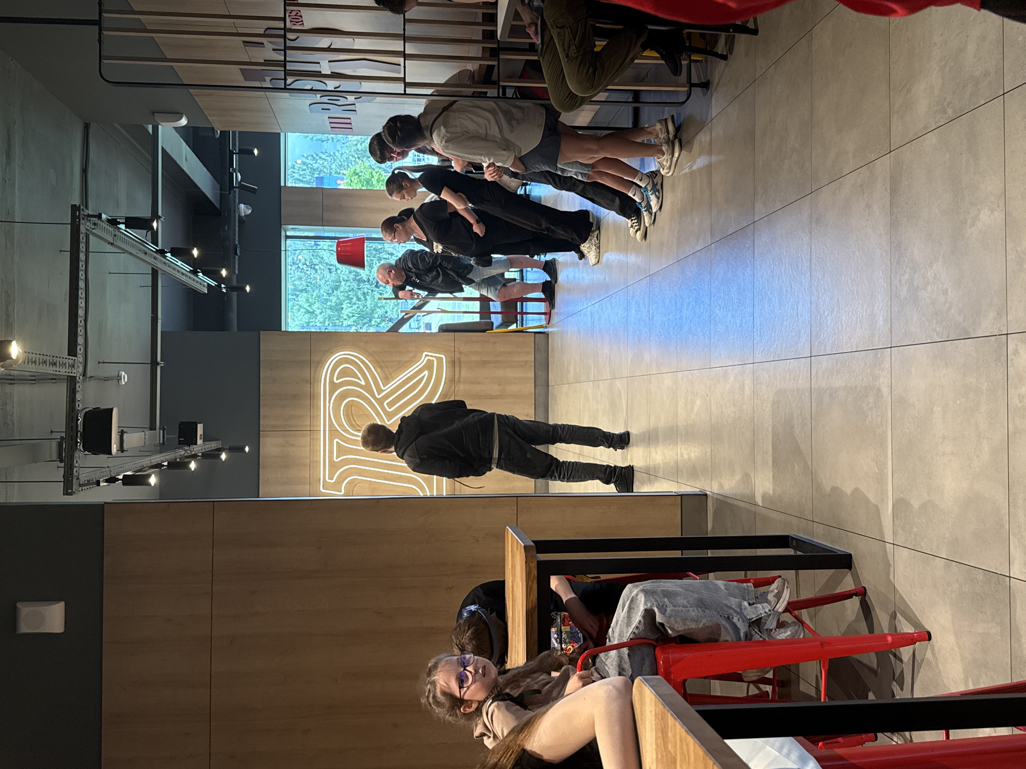 A group of people are standing and sitting in what appears to be a fast-food restaurant or cafe. In the foreground, a young girl with glasses is seated at a table, looking towards the camera. To her right, other individuals are standing in a line, possibly waiting to order or for their food. The environment features modern interior design with wooden panels, exposed ceiling elements with lighting fixtures, and tiled flooring. A large neon sign with the letter "R" is visible on the wall. The context of Izhevsk, Russia is not ascertainable from the visual information provided in the image.
