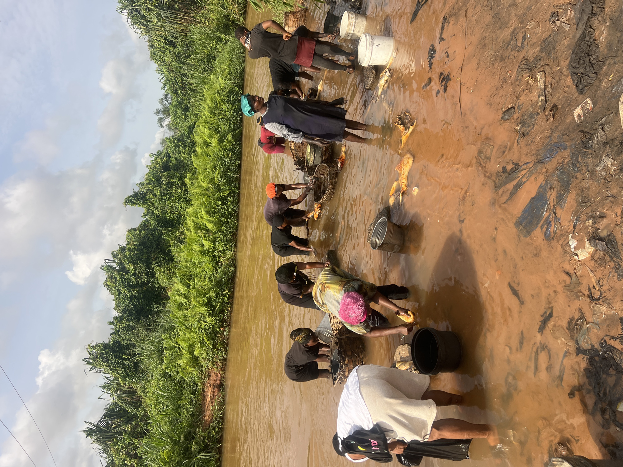 A medium, outdoor shot shows a group of people standing in a muddy river in Obosi, Nigeria. There are nine people, most of whom are women, standing in the reddish-brown river, some up to their knees. They are congregated in a line, facing the same direction and appear to be working or cleaning something. Each person has a basket or bucket nearby. The riverbank is littered with dark trash. Lush green vegetation lines the opposite bank, extending toward a cloudy sky.