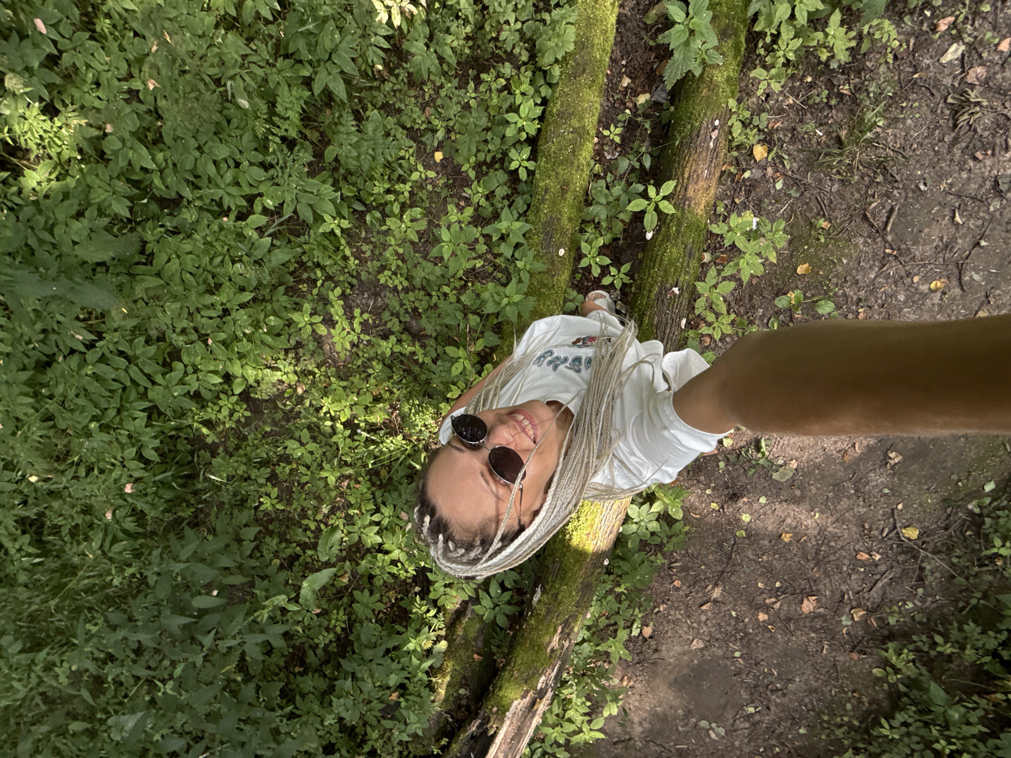 The media file is a high-angle, full-body shot of a young person with blonde braided hair, wearing sunglasses and a white t-shirt, standing on a fallen tree trunk in a wooded area. The person is smiling and looking up at the camera, appearing joyful and relaxed. The setting is a natural environment with abundant green foliage, moss-covered tree trunks, and a dirt path. The lighting suggests it is daytime, possibly late morning or early afternoon, with sunlight filtering through the trees. The overall mood is cheerful and serene, capturing a moment of enjoyment in nature. The location is identified as Rusanovo, Russia. No visible text is present in the image.